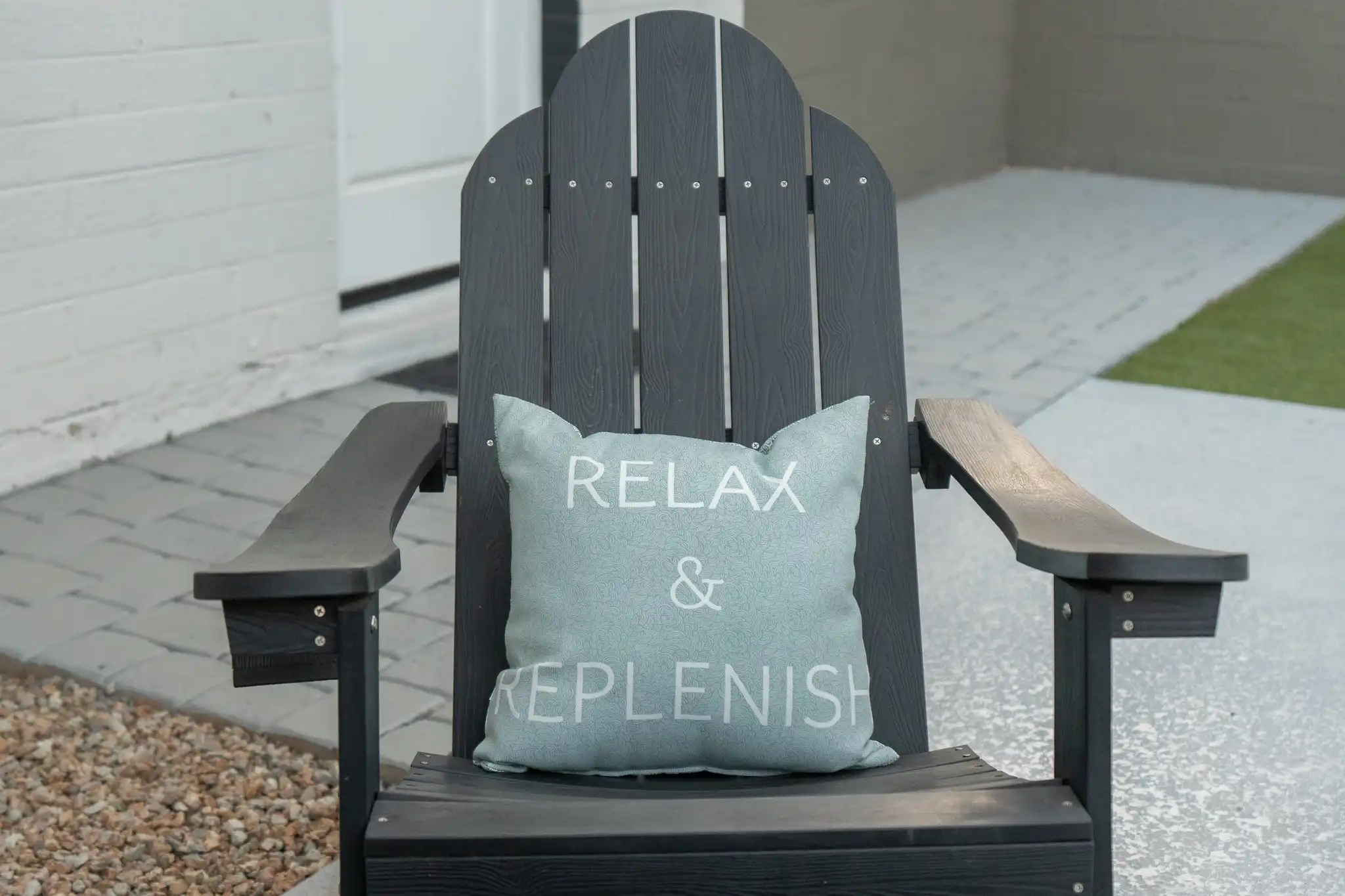 A dark gray Adirondack chair on a patio holds a light blue throw pillow inscribed with the words 'RELAX & REPLENISH'.