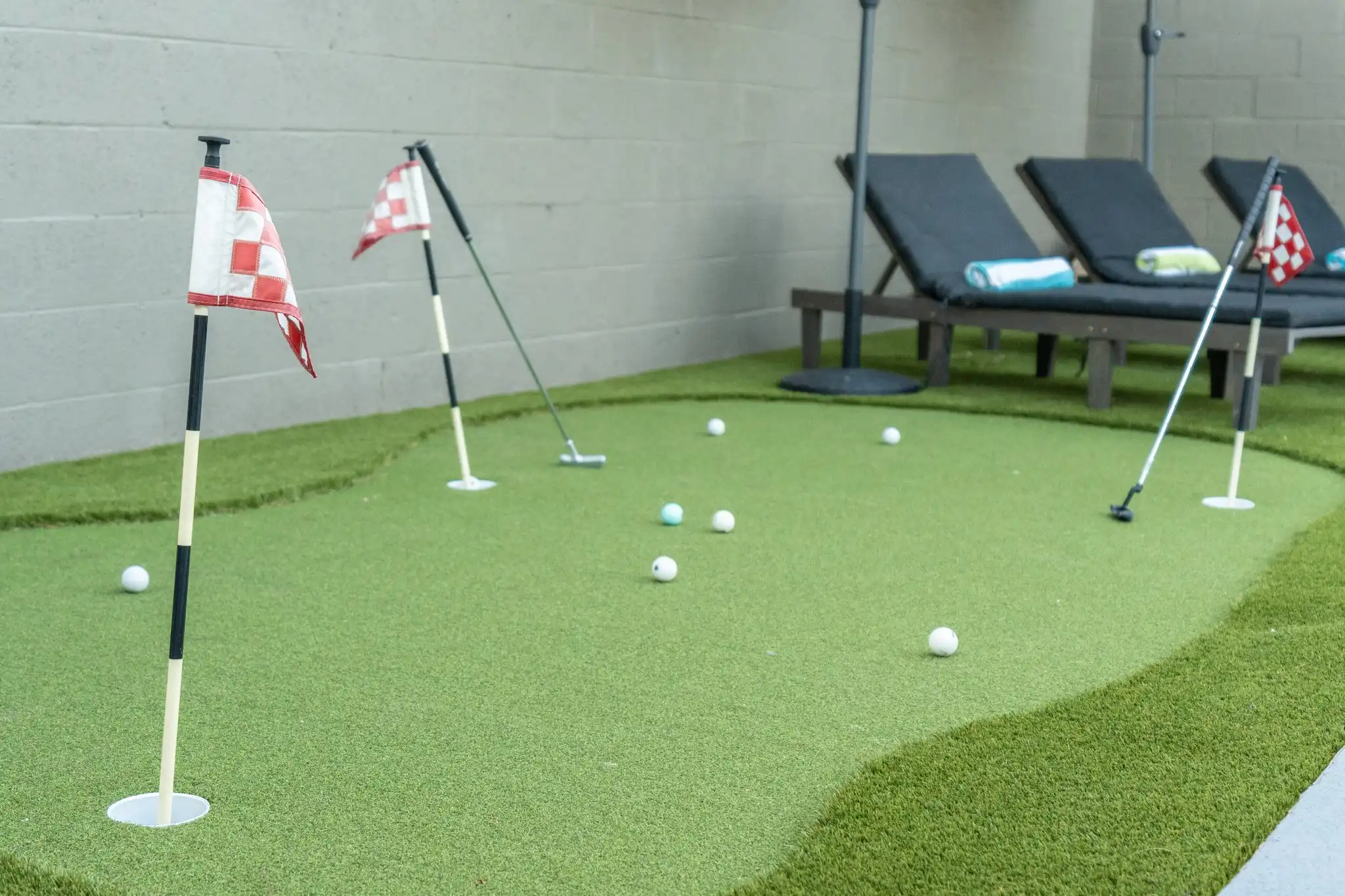 An outdoor miniature golf putting green on artificial turf, showing several golf balls, a putter, and two checkered flags. Lounge chairs are visible behind the green.