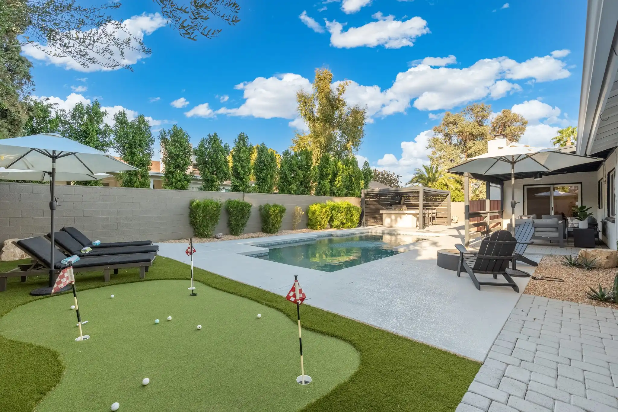 A sunny backyard with a rectangular pool, a putting green with flags, and several lounge areas. The space is landscaped with hedges and trees under a blue sky.