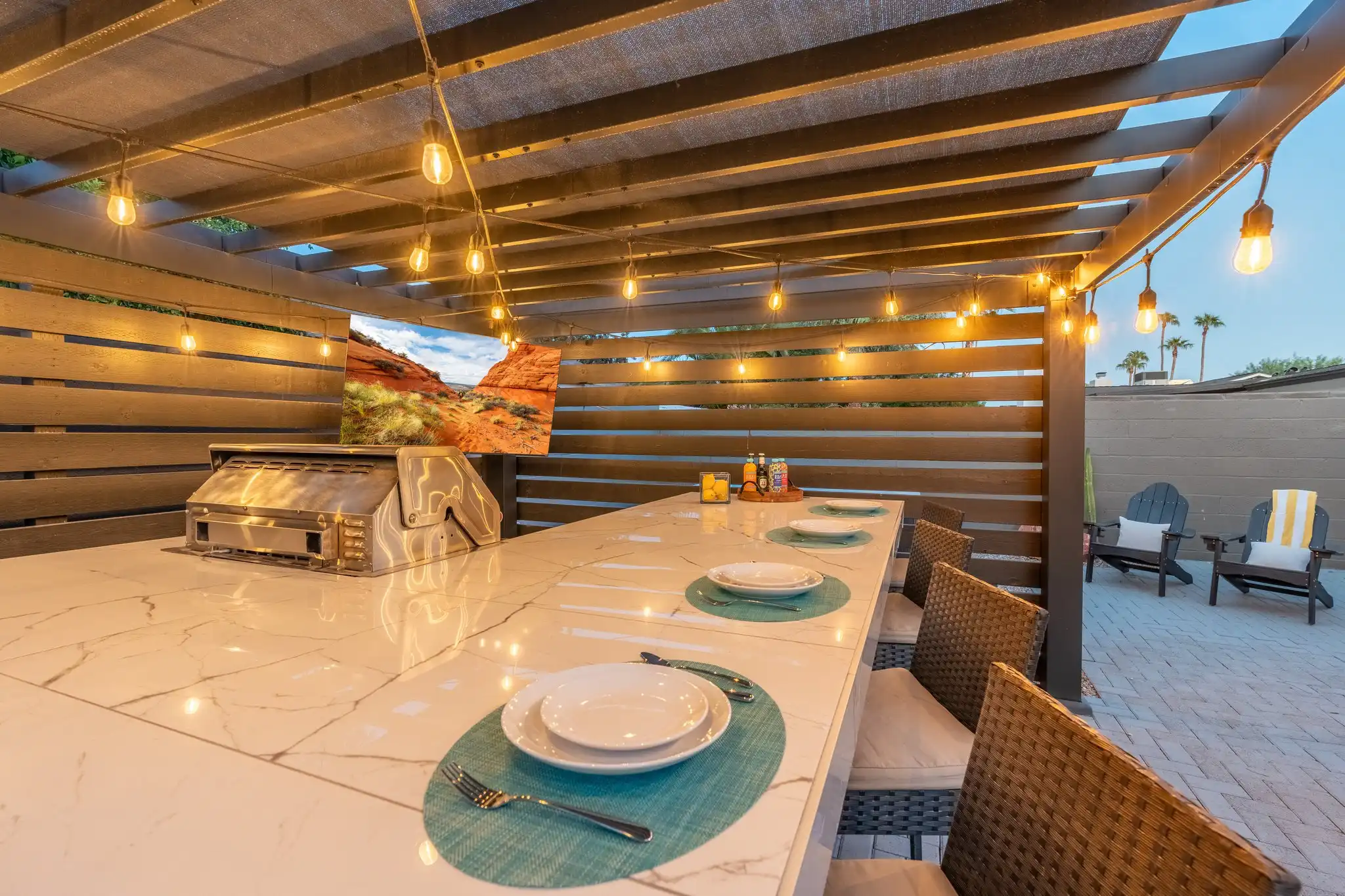 An outdoor kitchen bar beneath a pergola, featuring a white marble counter, place settings, and wicker stools. String lights illuminate the built-in grill and a TV display.