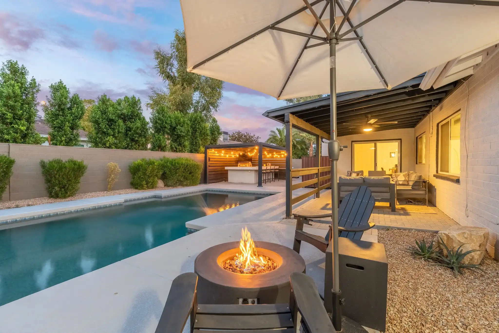 A modern residential backyard features a fire pit, a long swimming pool, and multiple covered seating areas, illuminated by string lights at twilight.