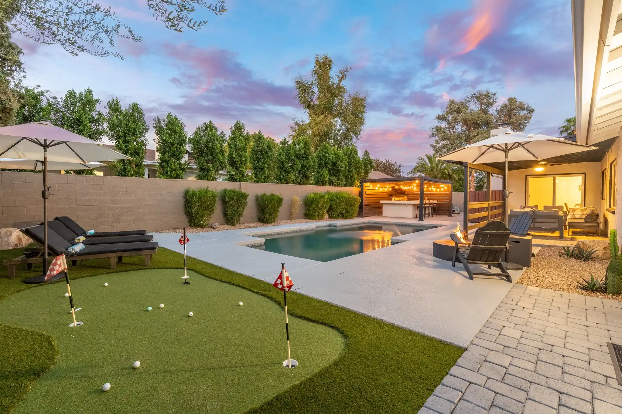 A luxurious backyard features a swimming pool, mini putting green, and an illuminated outdoor kitchen and patio area under a colorful sunset sky.