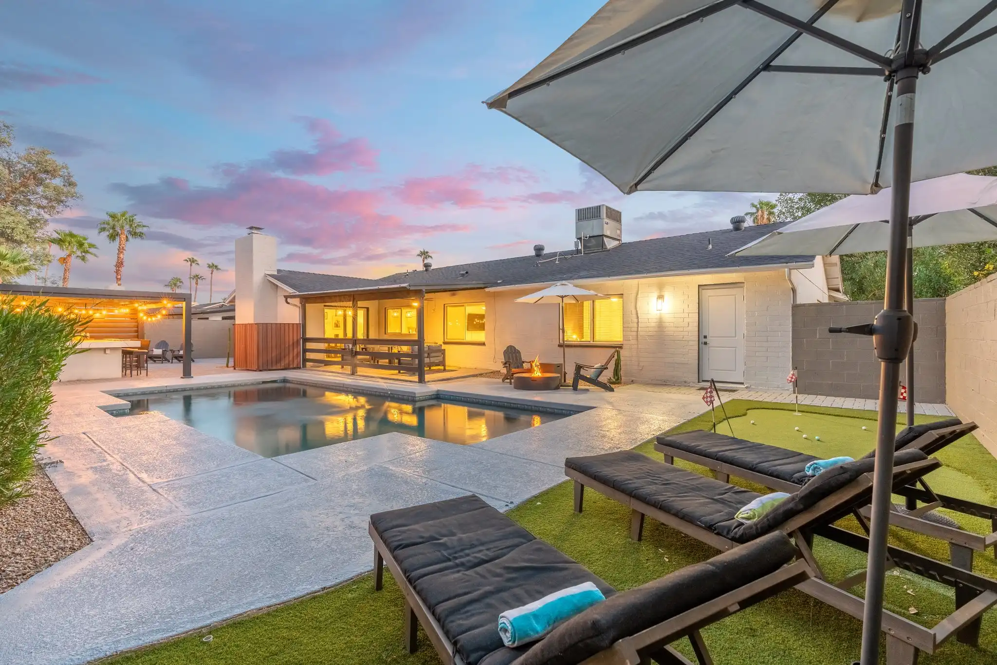 A luxurious backyard pool and patio at sunset featuring a fire pit, several lounge chairs, a covered outdoor bar, and a small putting green.