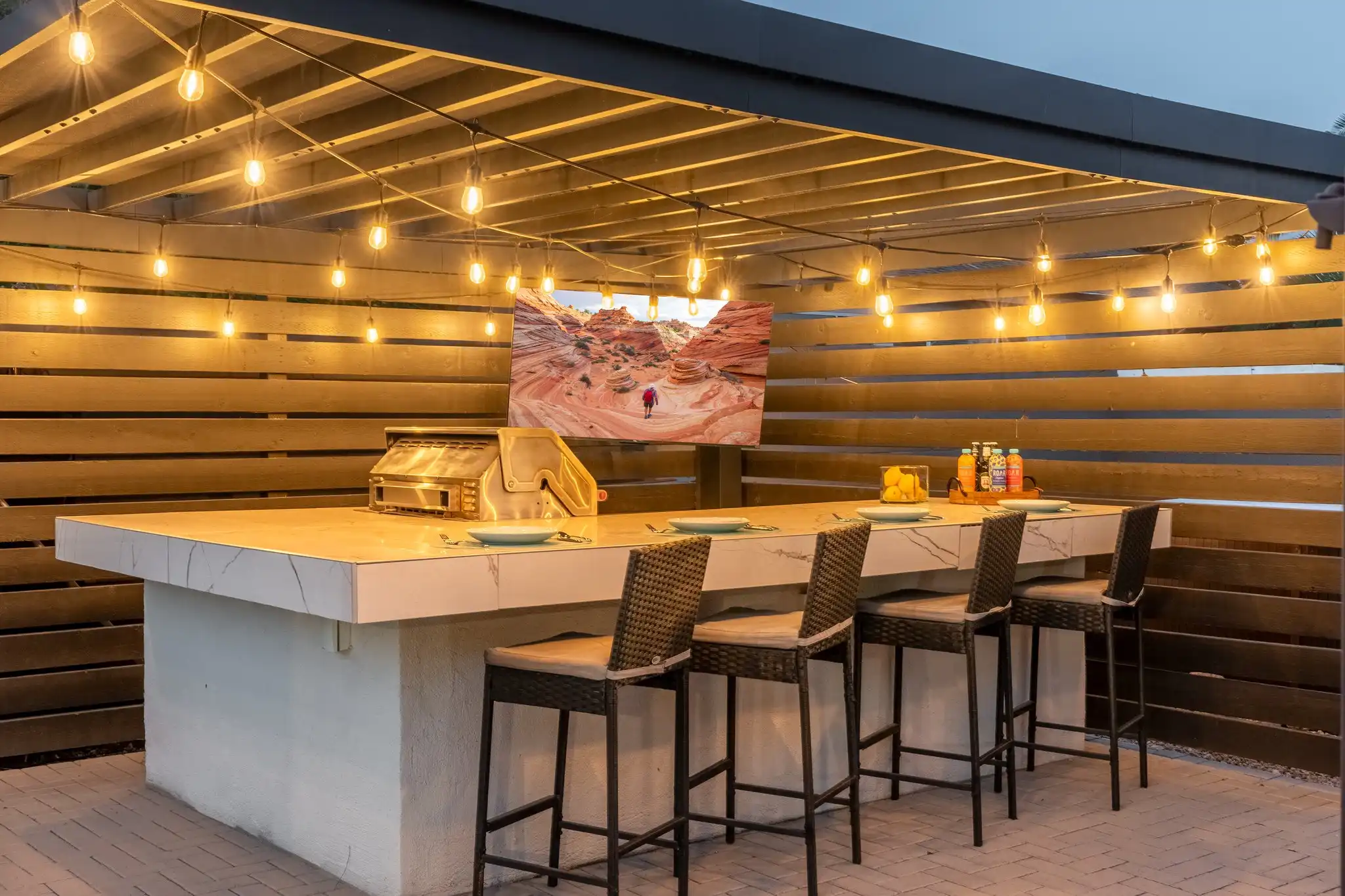 A stylish outdoor bar area with a white counter, four high stools, and a grill. String lights illuminate the space and a TV displays a desert scene.