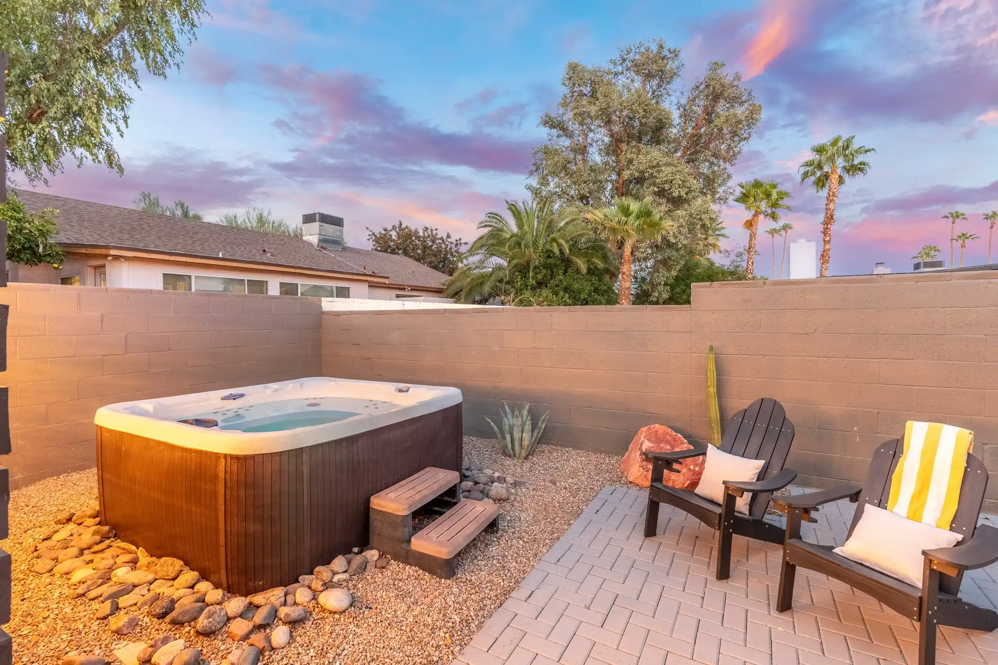 An outdoor hot tub and two Adirondack chairs situated in a private, desert-style backyard under a dramatic pink and blue sunset sky.