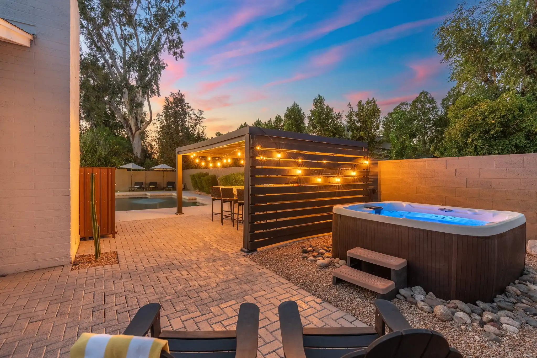 A twilight view of a resort-style backyard patio featuring a glowing hot tub, a covered pergola with string lights, and a paved area leading to a pool and loungers.