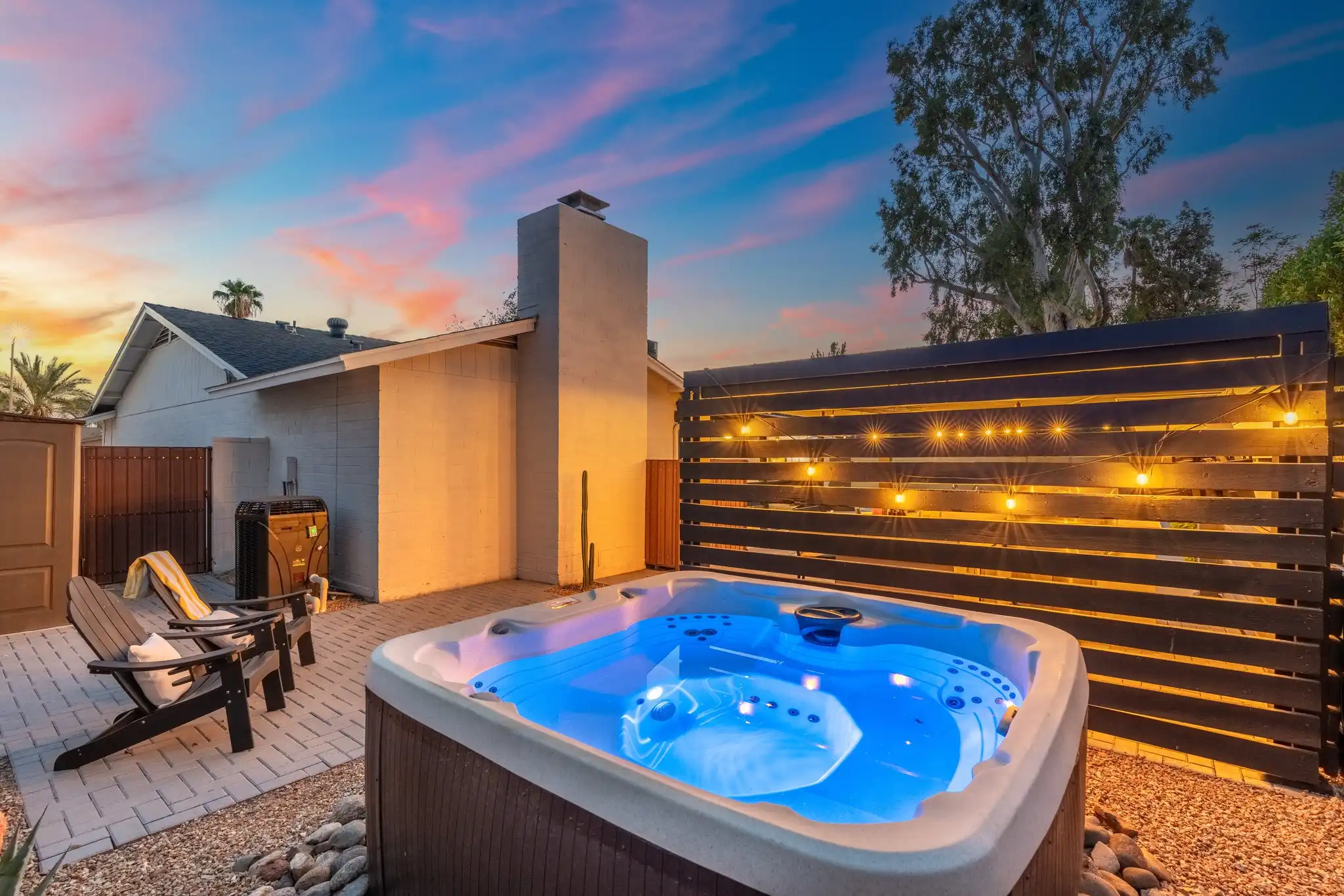 A modern hot tub glows blue on a stone patio at sunset. String lights illuminate the nearby wooden fence and two Adirondack chairs.