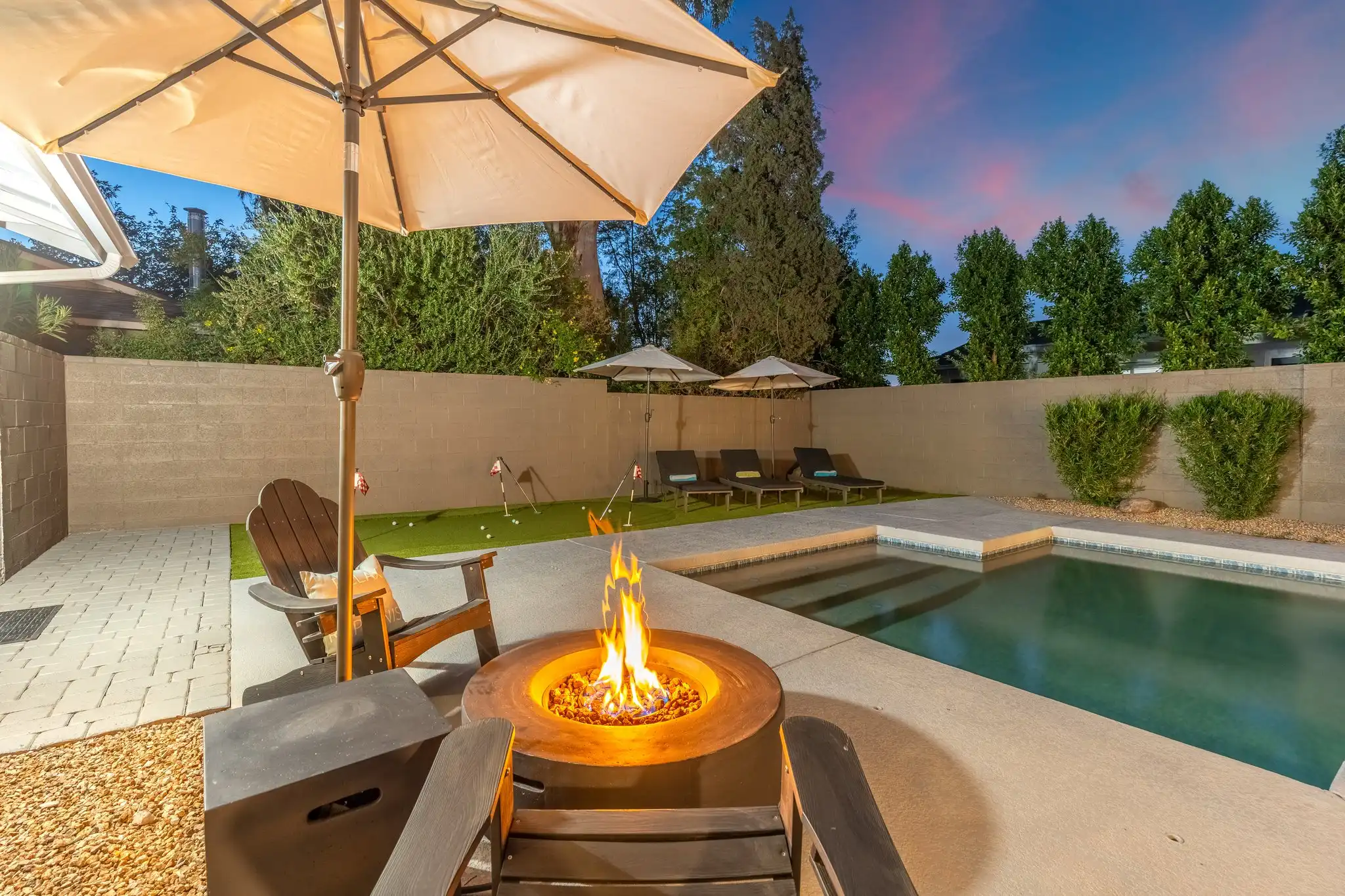 A glowing fire pit and chairs sit beside a swimming pool at twilight, with a putting green and lounge chairs visible against the backdrop of trees.