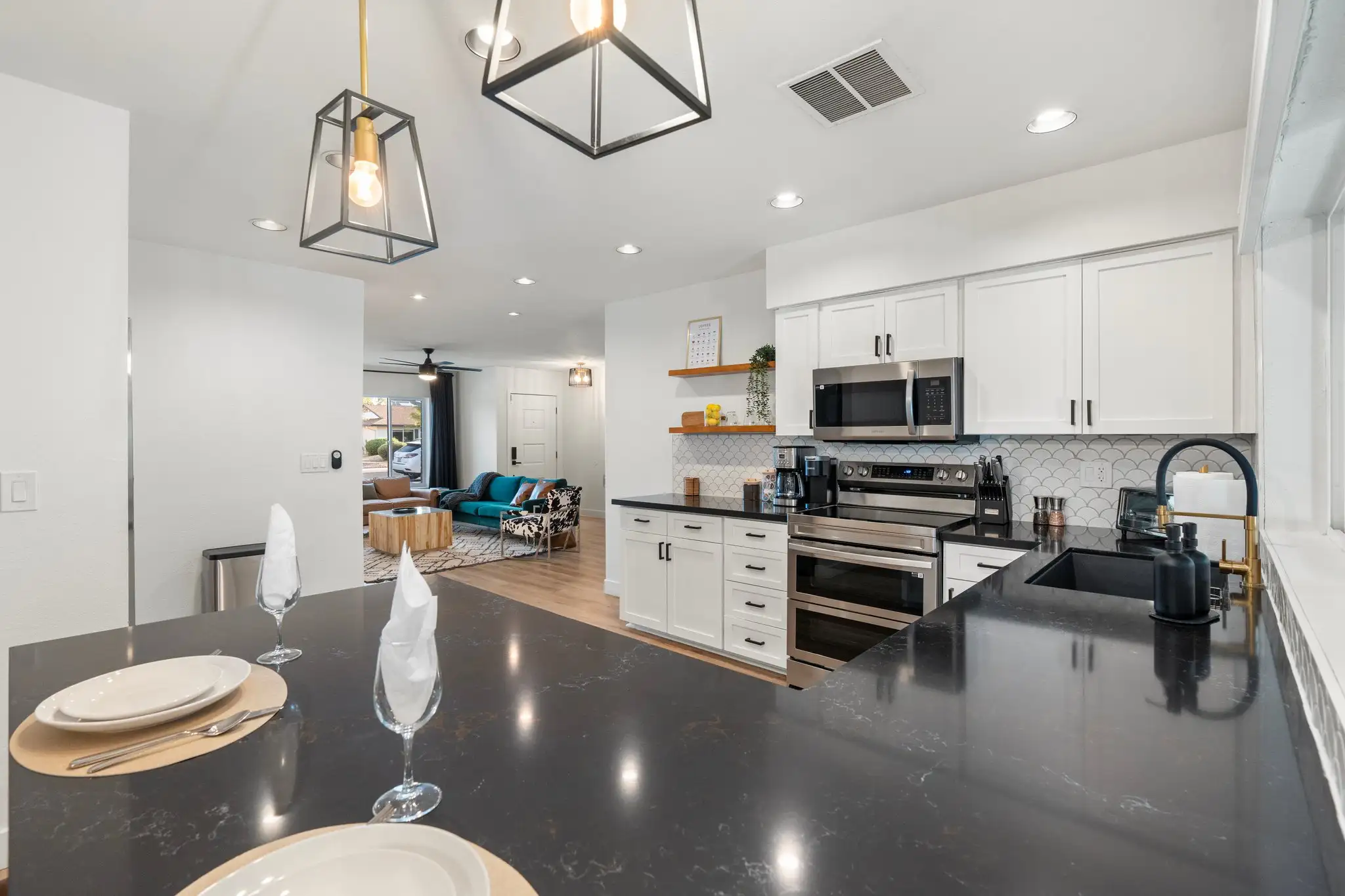 A modern, open kitchen with black counters and white cabinets. The island is set with plates, looking toward a living room furnished with a teal couch.