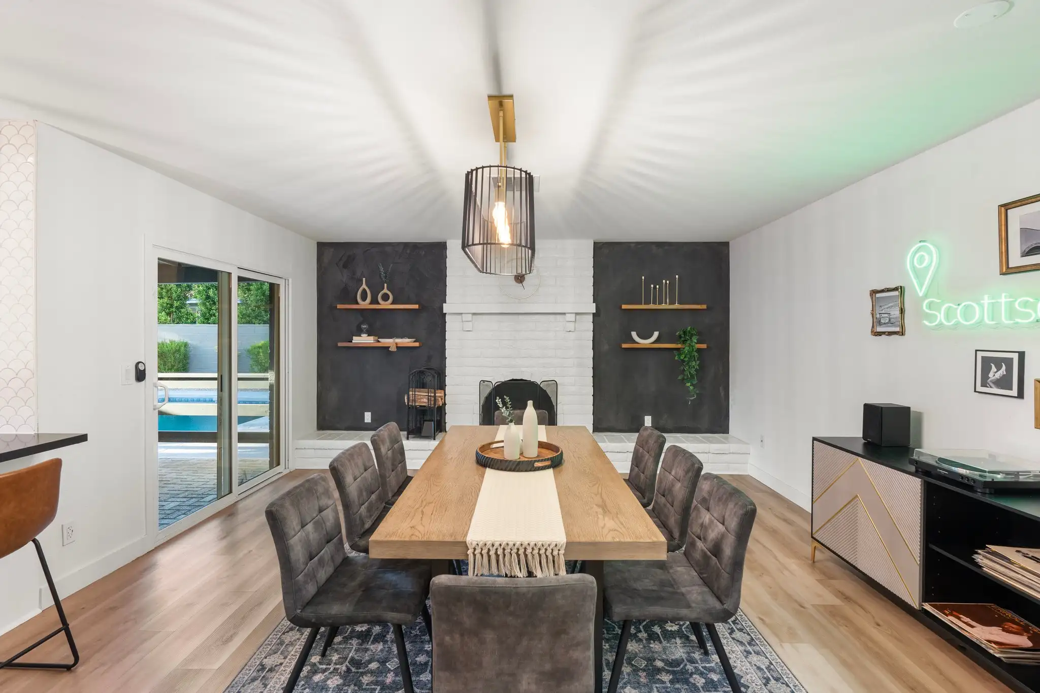 A modern dining room featuring a wood table with gray upholstered chairs. The space includes a white brick fireplace, dark accent walls, and sliding doors opening to a backyard pool.