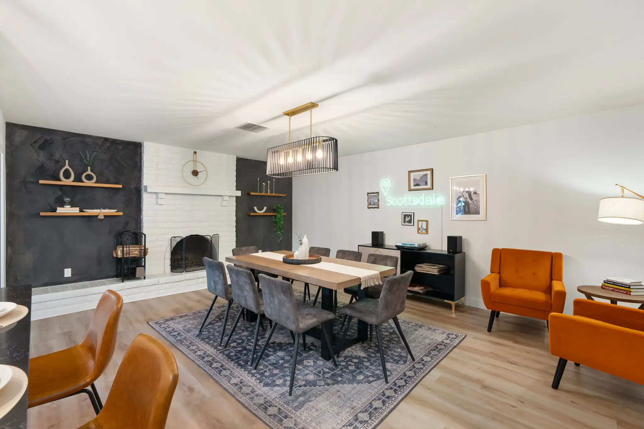 A stylish dining room featuring a long wood table and eight gray chairs on a rug. Decor includes a white brick fireplace, dark accent walls, and a "Scottsdale" neon sign above a black sideboard.