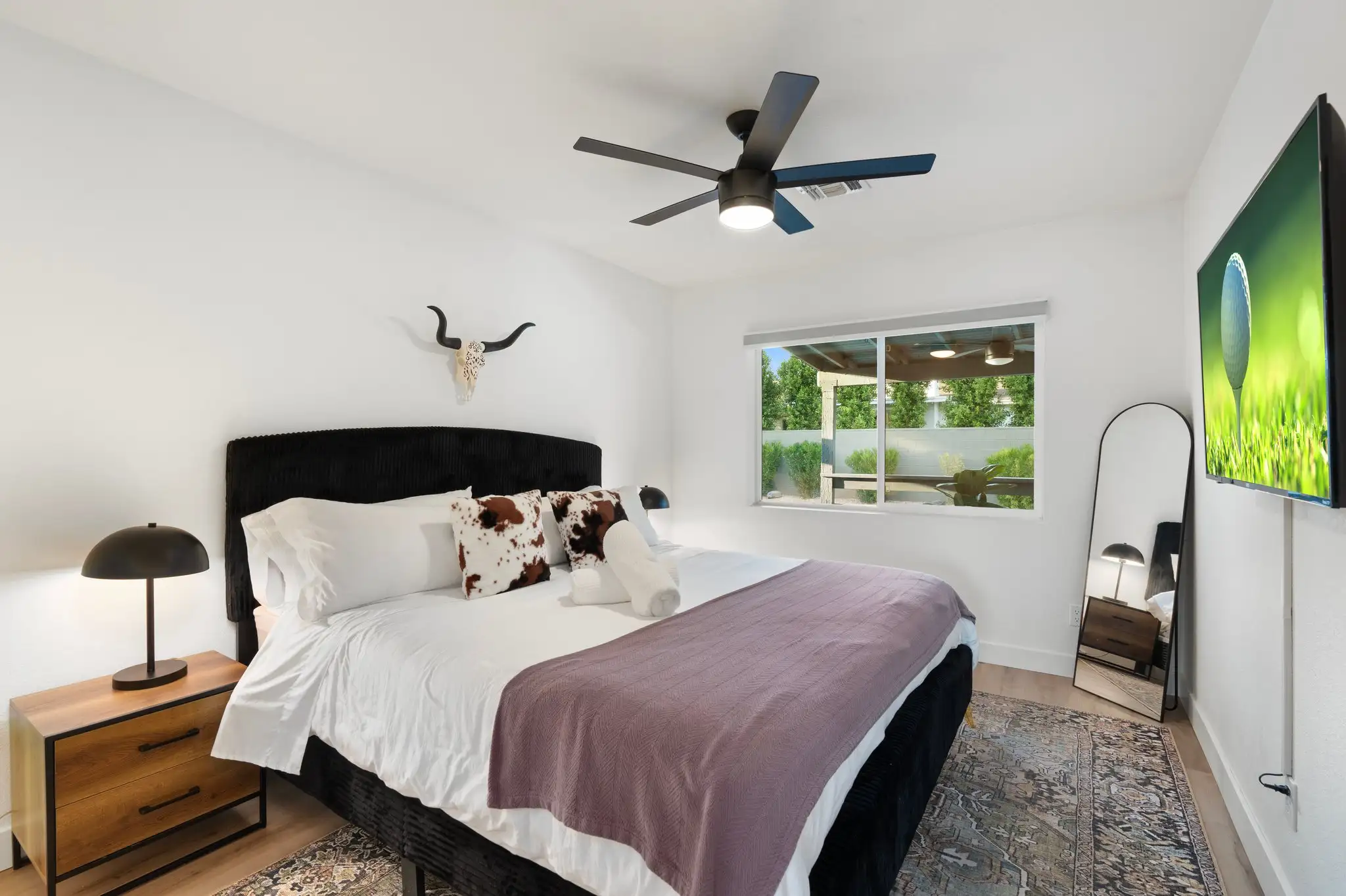 A bright white bedroom with a western aesthetic, featuring a black velvet bed, cowhide pillows, and a steer skull decoration. The room has a modern ceiling fan, a large floor mirror, and a window overlooking a patio.