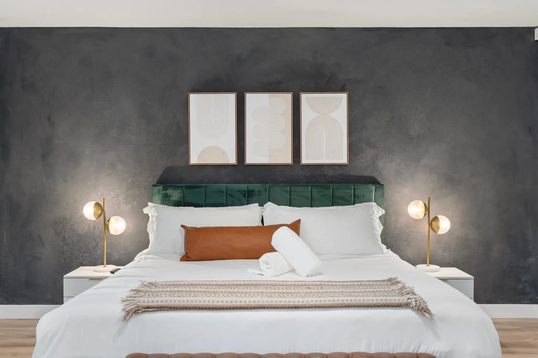 A sophisticated bedroom with a dark gray wall, green velvet headboard, and white bed linens, accented by gold globe lamps and abstract art.