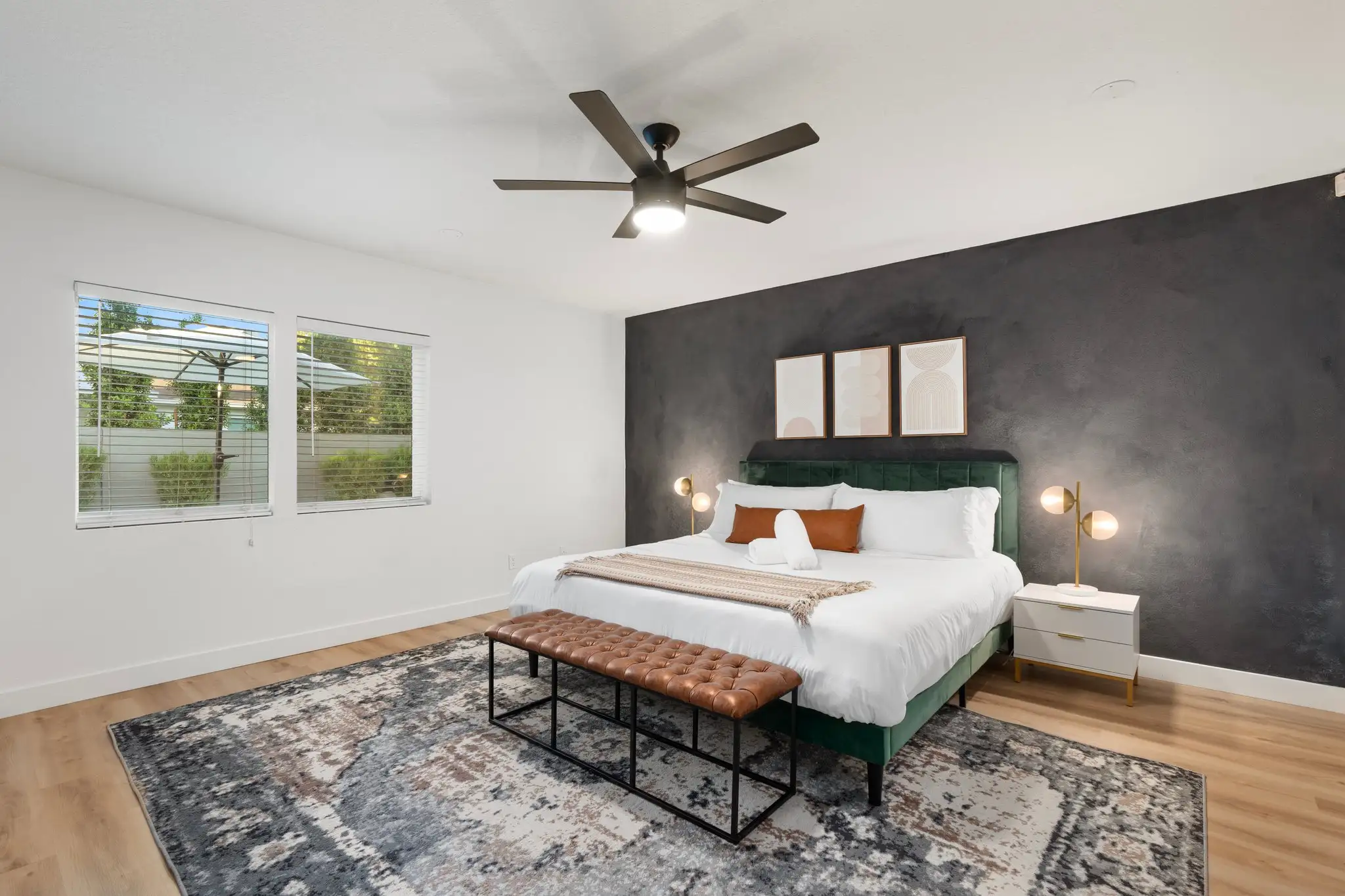 A stylish bedroom with a dark gray accent wall, a green velvet bed, and white linens. A tufted brown leather bench rests on a large abstract rug.