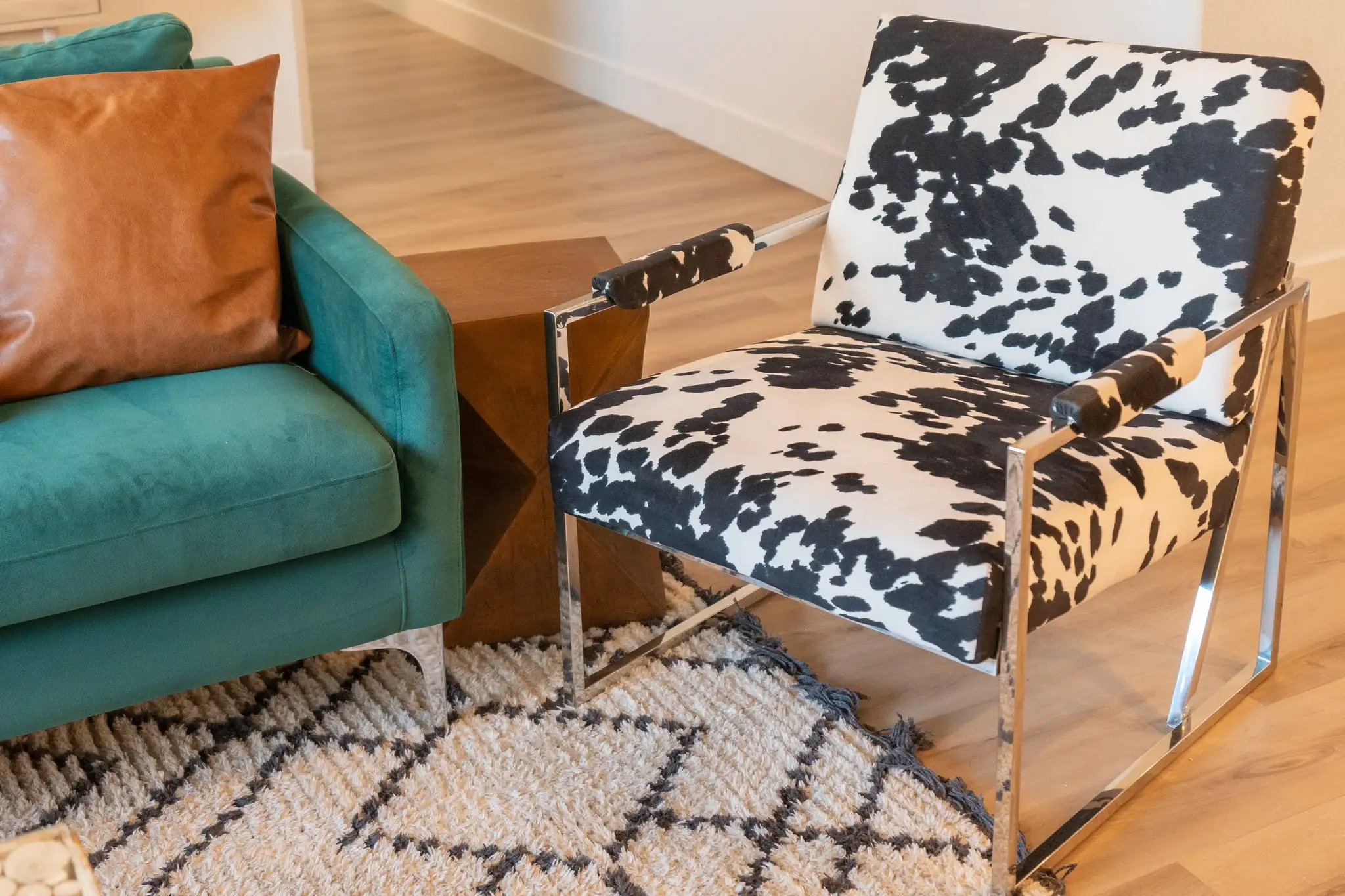 A stylish contrast of textures: a black and white cow print armchair stands next to a teal velvet sofa and a wood side table over a fuzzy rug.