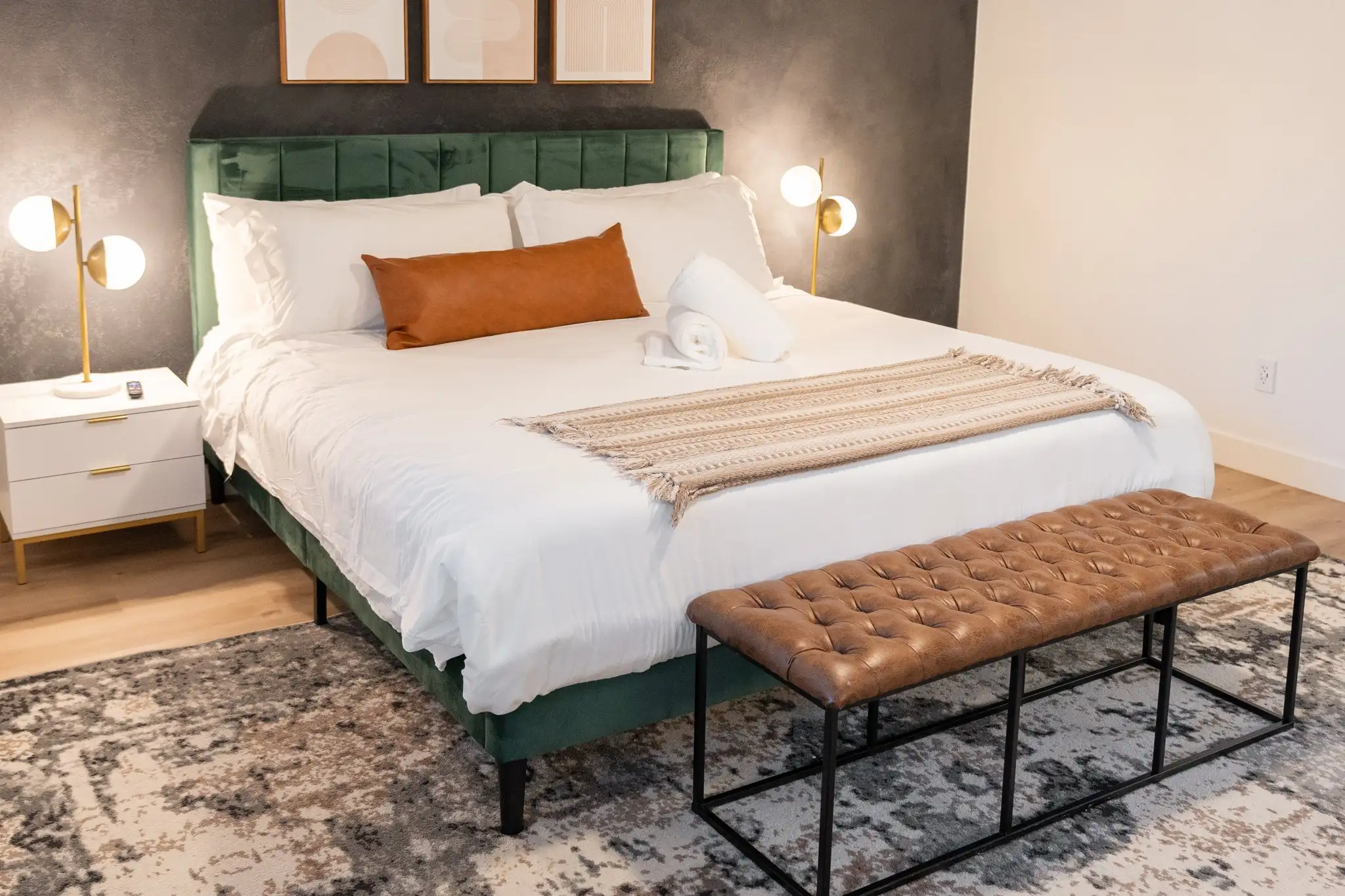 A modern king bed with a green velvet headboard, white sheets, and a rust-colored pillow. A tufted leather bench rests at the foot, on an abstract area rug.