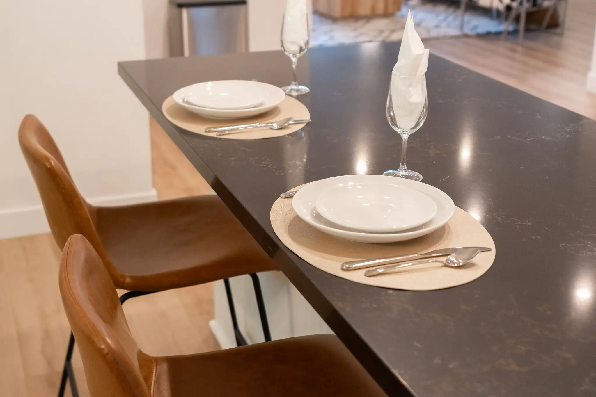 Two brown leather counter stools sit next to a dark gray quartz kitchen island, which is formally set with two placemats, white plates, and wine glasses.