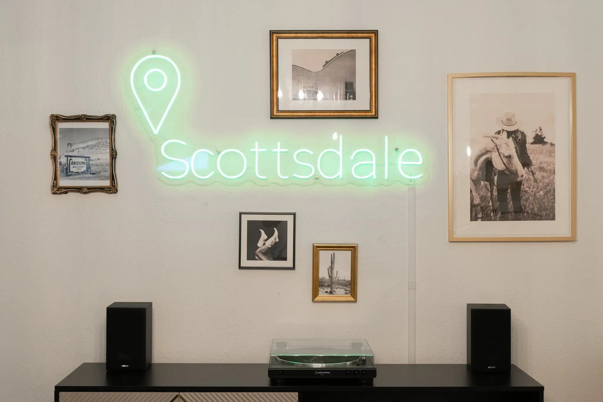 A white wall features a green neon sign that says "Scottsdale," surrounded by framed black-and-white photos of the desert, above a record player.