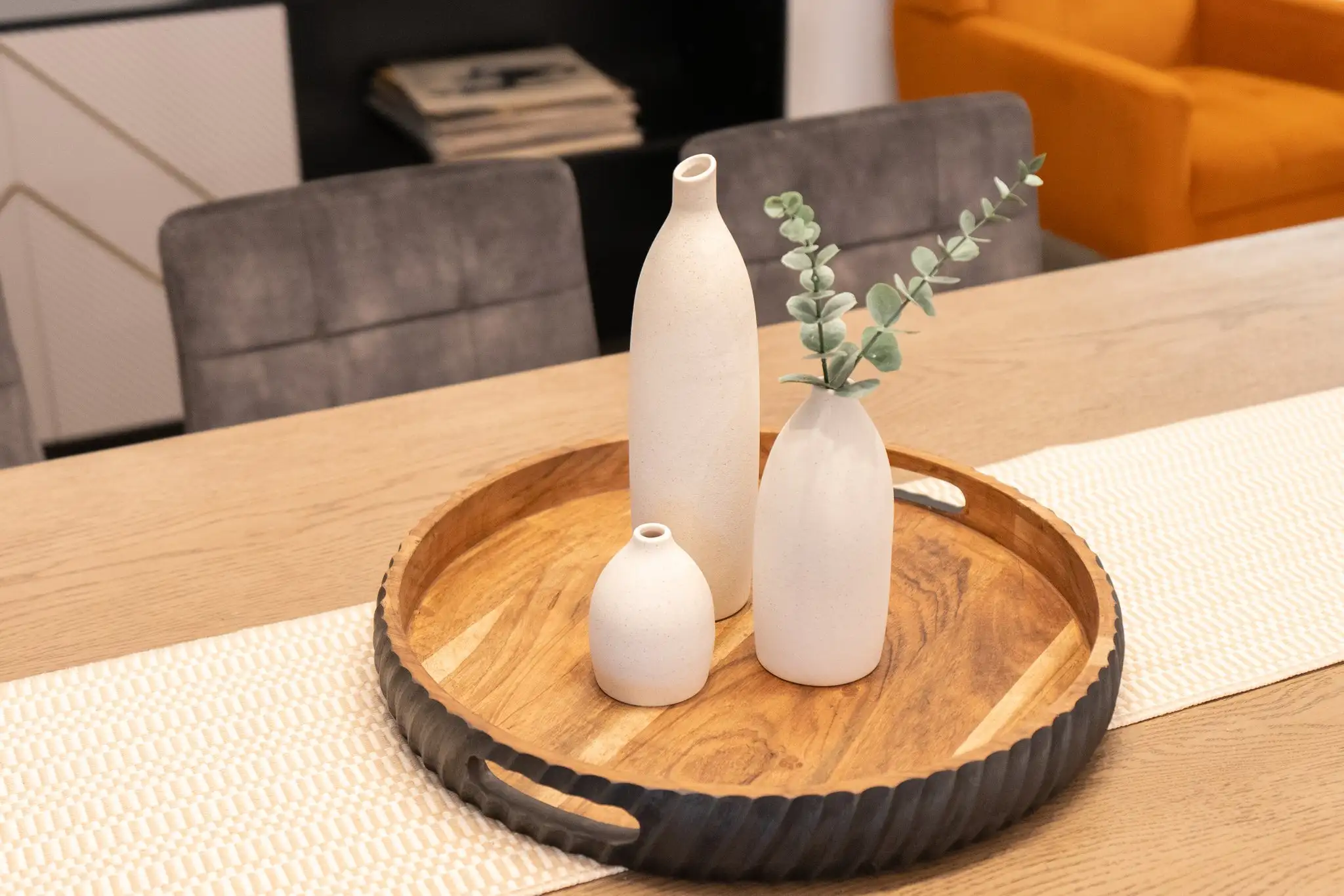 Three modern white textured vases, one holding eucalyptus leaves, are displayed on a round wooden serving tray on a light wood table.