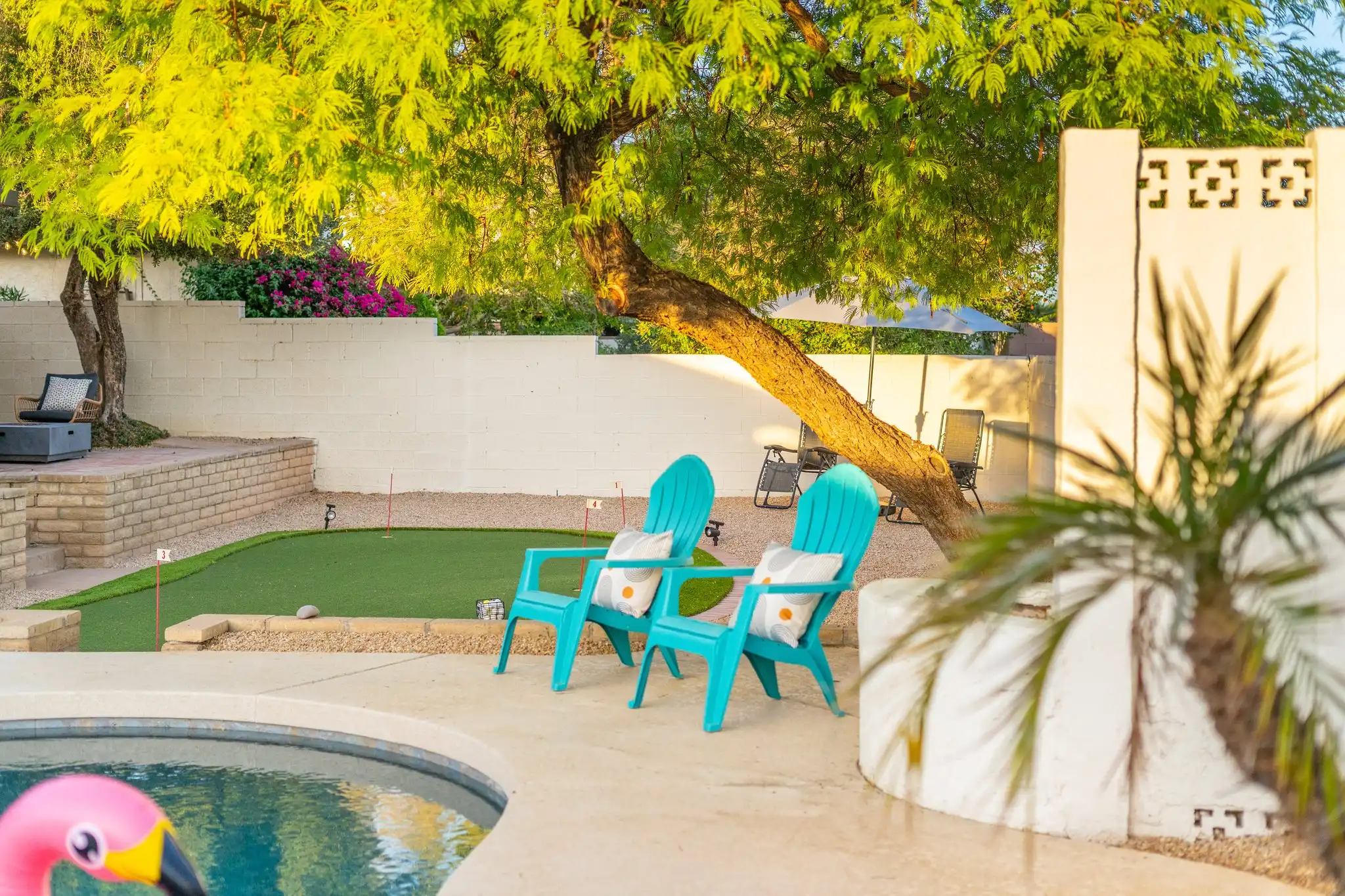Two bright teal Adirondack chairs rest on the concrete patio near a pool, shaded by a large tree. Behind them is a manicured putting green and white landscape walls.