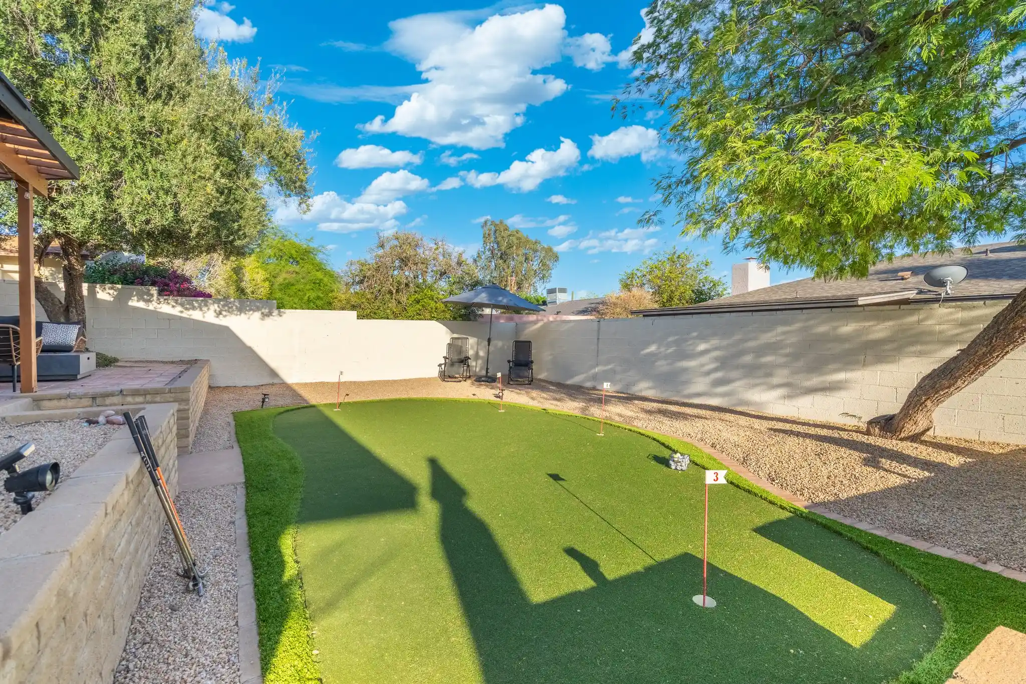 A sunny residential backyard with a bright blue sky, featuring a curved artificial turf putting green, surrounded by gravel and trees.