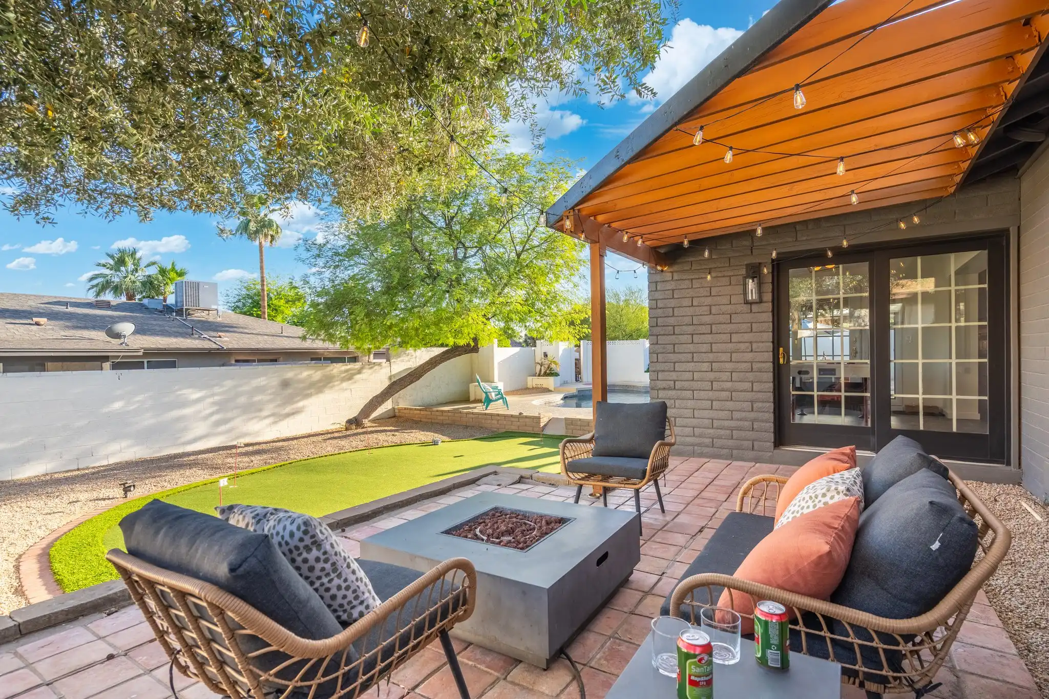 An inviting outdoor patio features woven seating, a central fire pit, and string lights above a brick floor. The backyard space includes a small putting green under a bright sky.