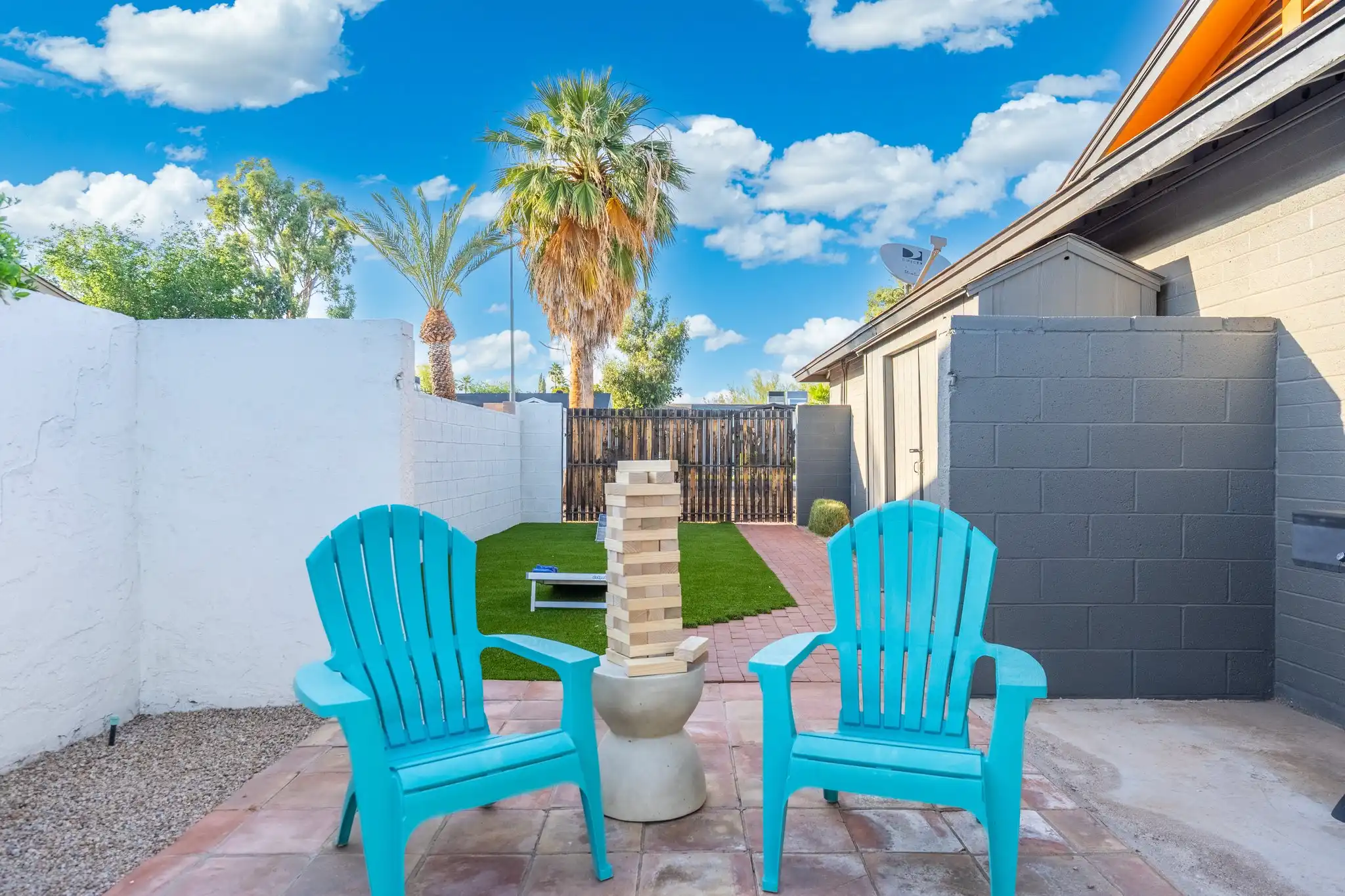 Two bright turquoise Adirondack chairs frame a giant Jenga game set up on a patio, leading to an artificial turf area for backyard games under a sunny sky.