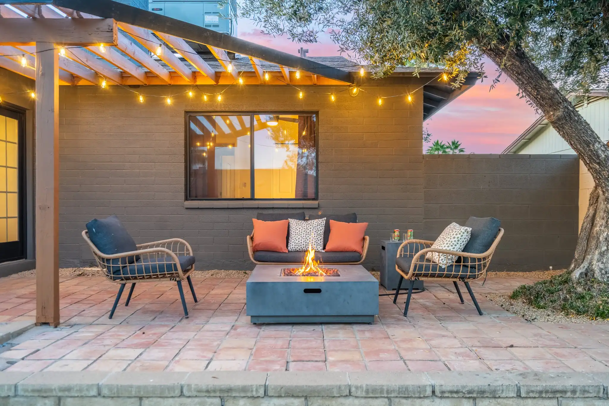 A cozy patio at twilight featuring a lit fire pit surrounded by wicker seating with dark cushions. A wooden pergola with string lights hangs over the area.