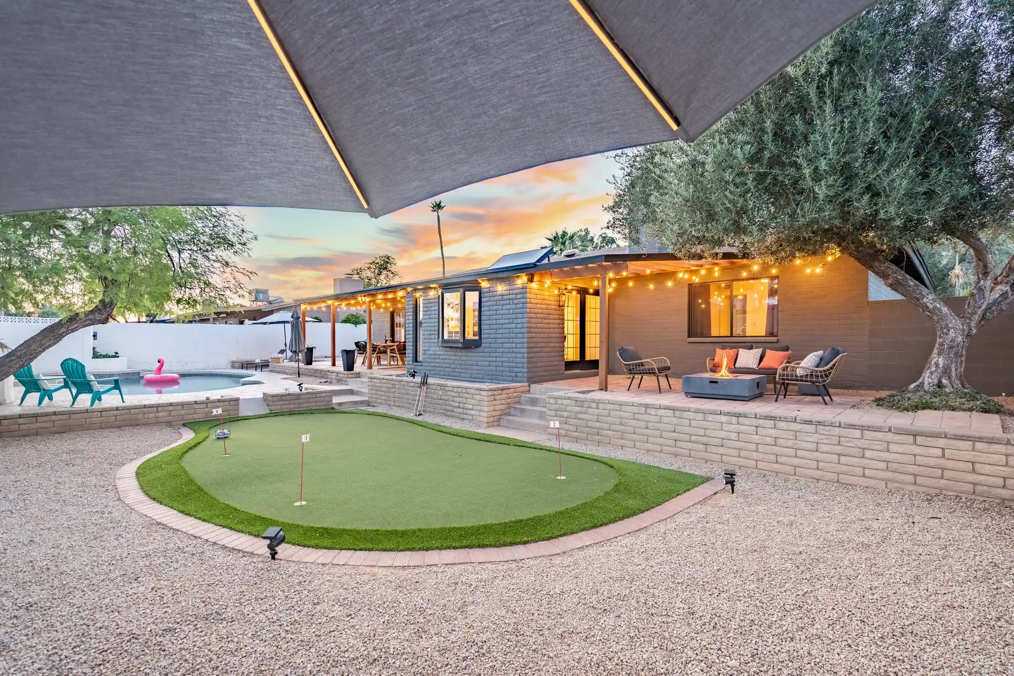 A twilight view of a modern residential backyard featuring a curved artificial turf putting green set in gravel. The raised patio includes string lights, a lit fire pit seating area, and a swimming pool.