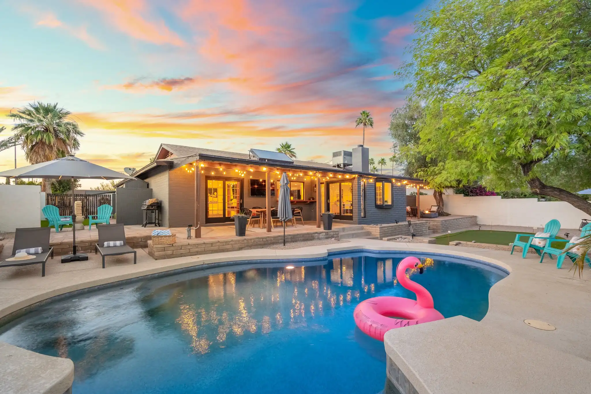 A modern backyard at sunset showing a house lit by string lights, a pool with a pink flamingo floatie, lounge chairs, and palm trees.