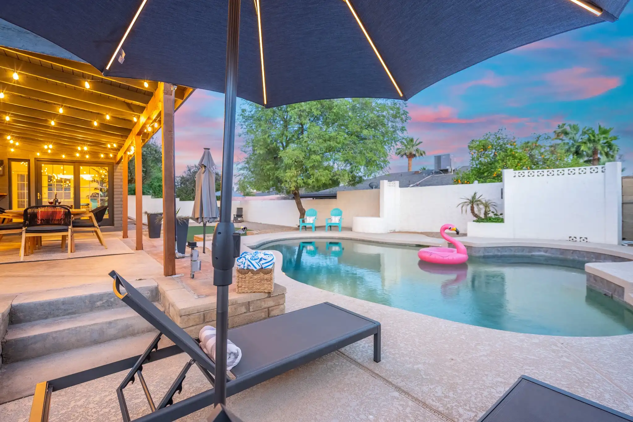 A resort-style backyard pool area at dusk. String lights glow on the covered patio near a dining area. A pink flamingo float is in the pool beside a lounge chair.