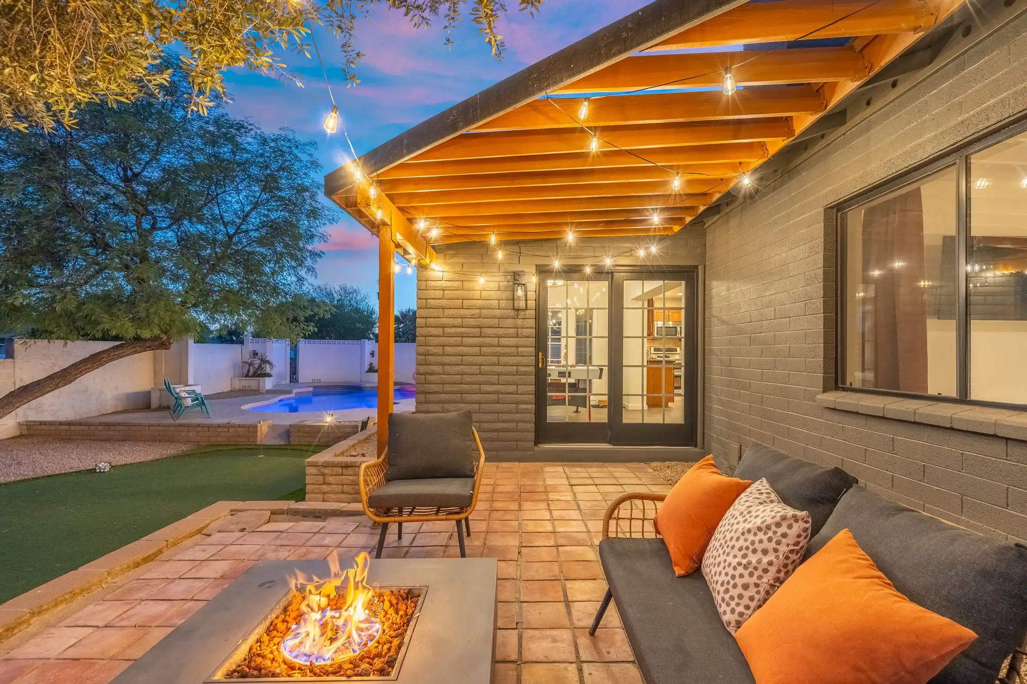 A modern backyard patio at twilight with a lit fire pit and dark outdoor seating. String lights illuminate the wooden pergola above the gray brick exterior wall.
