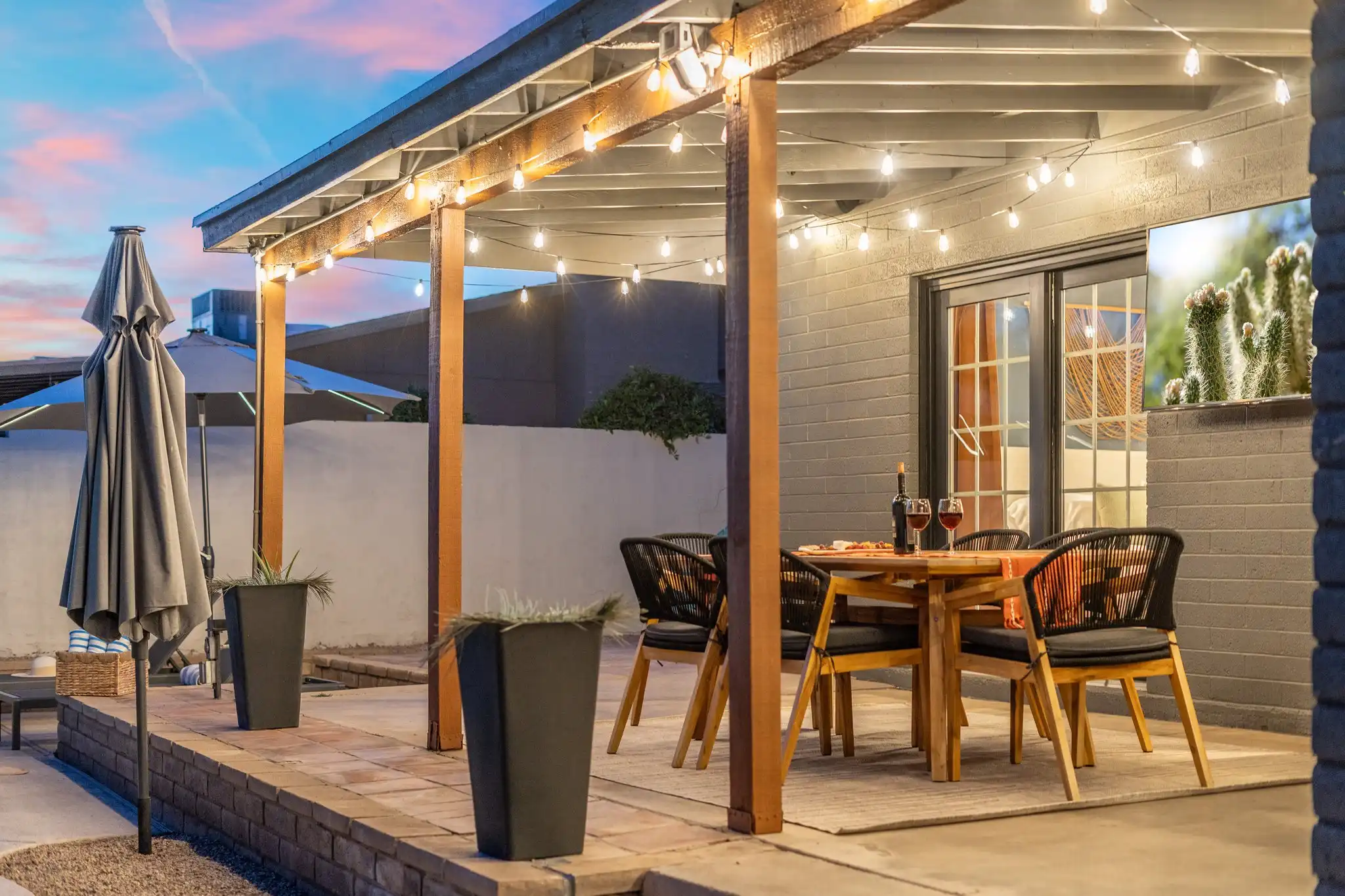 A covered outdoor patio at dusk, lit by string lights. A wooden dining table is set with wine and four woven chairs near sliding glass doors.