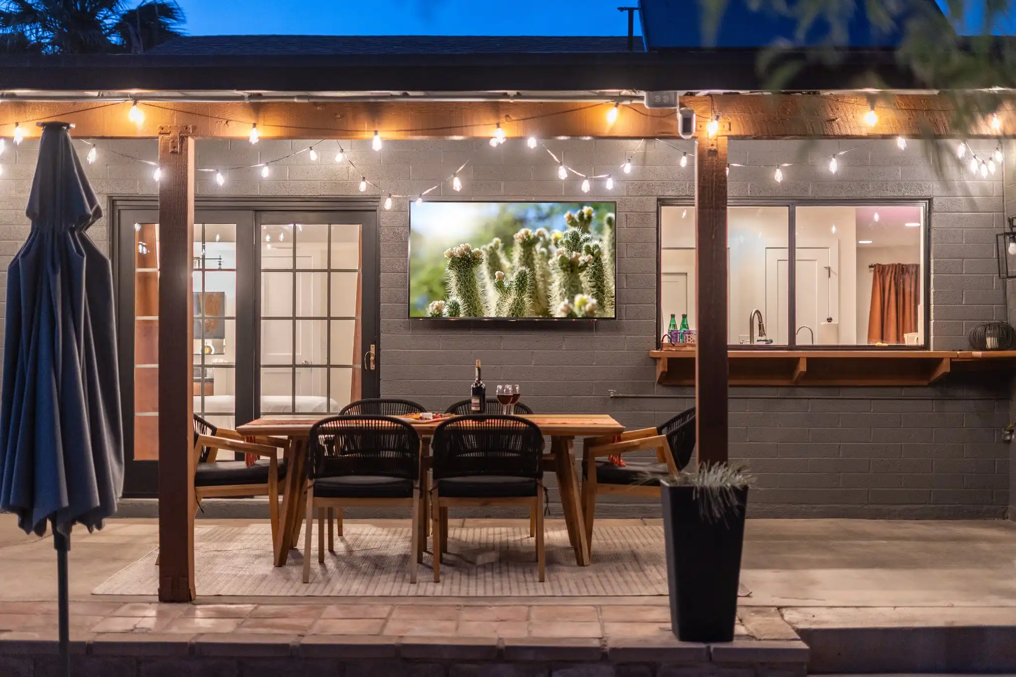An evening view of an outdoor patio with a dining set and string lights. A TV displaying a cactus is mounted on the dark brick wall.