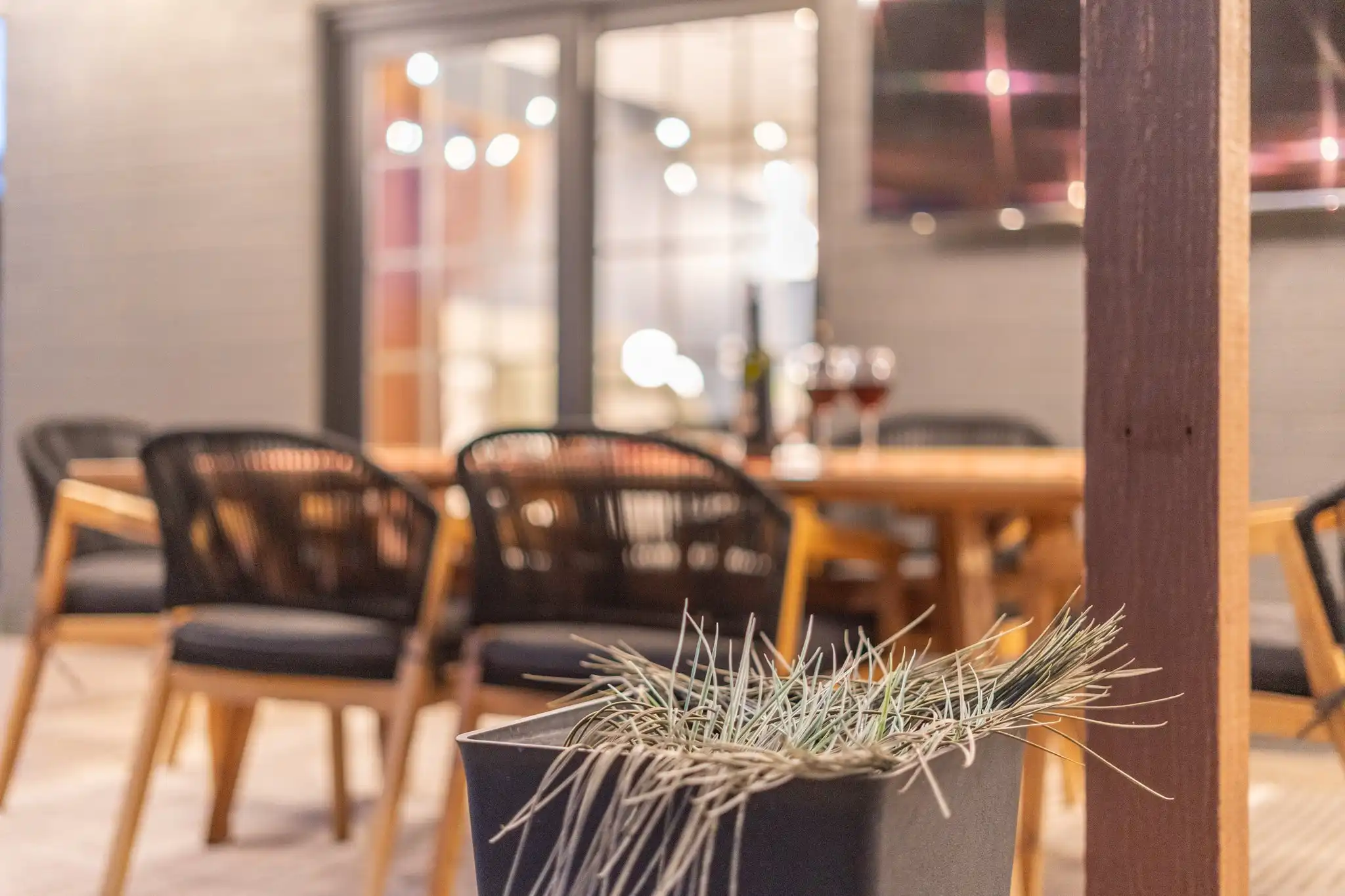 A modern exterior dining area with a wooden table, black chairs, and wine. Bright lights glow in the background behind glass doors.
