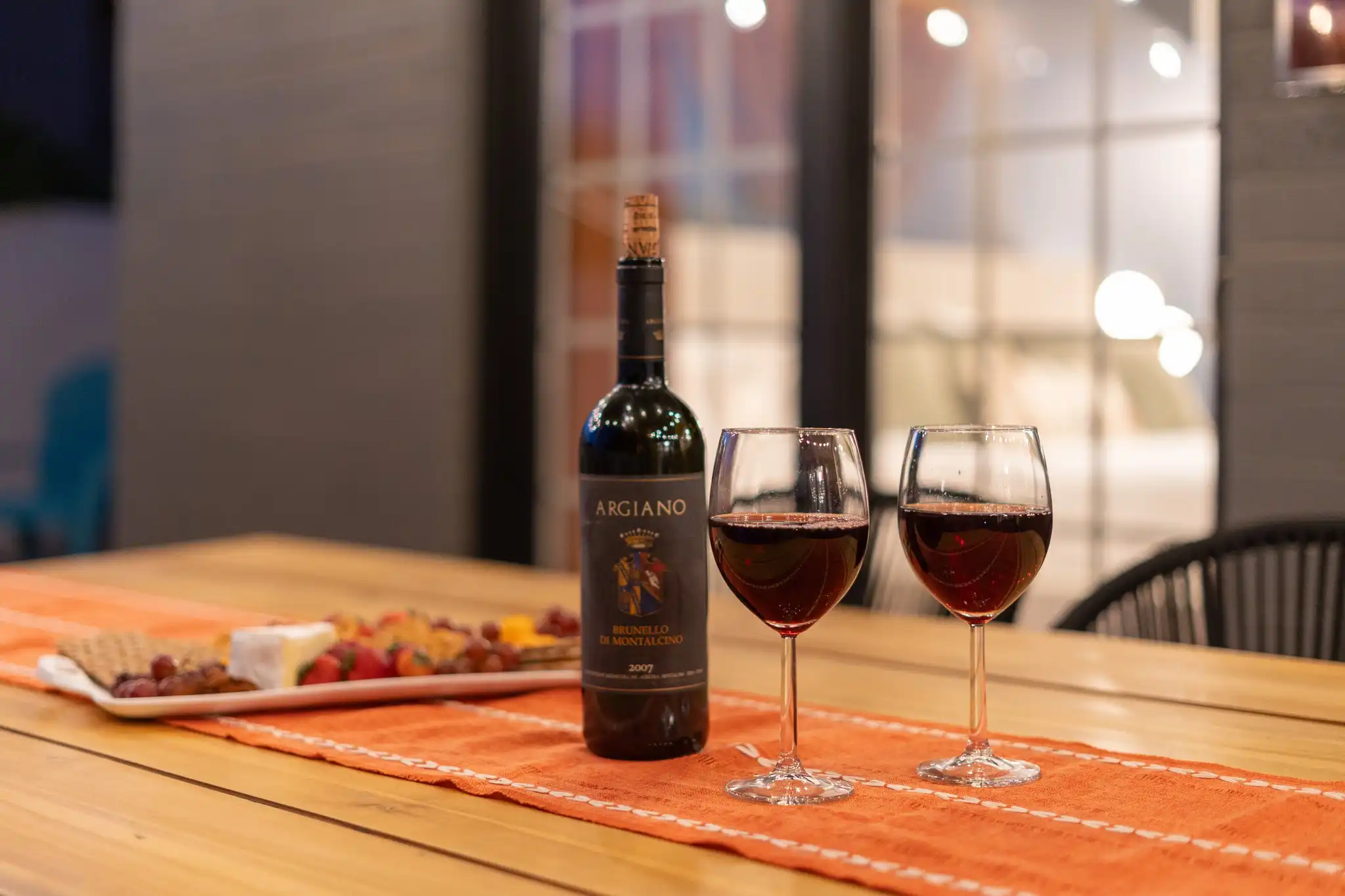 Two glasses of red wine and a bottle are placed on an outdoor patio table next to a platter of cheese, grapes, and crackers for an evening aperitif.
