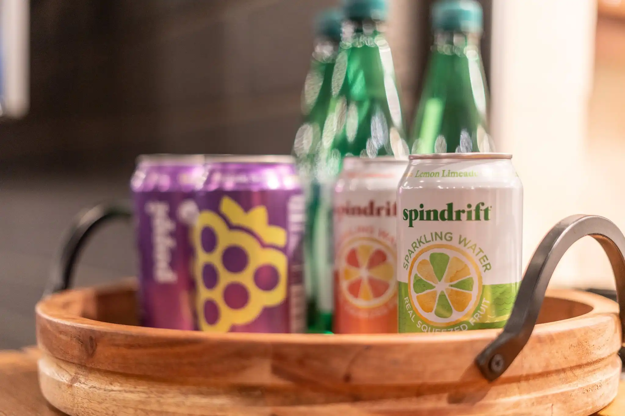 A wooden serving tray holds an assortment of canned and bottled beverages, including sparkling water and a purple can with a grape logo.