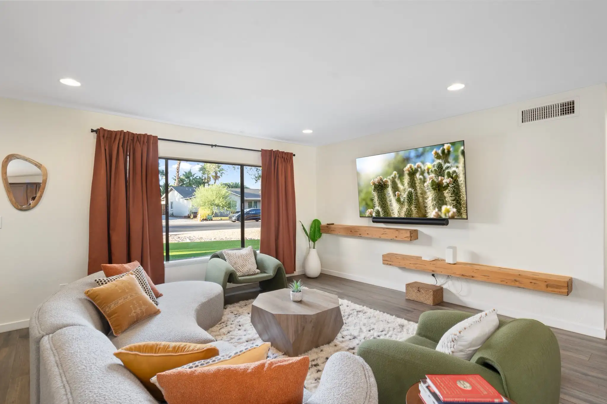 A spacious living room featuring a curved gray sofa and two green chairs arranged around a wooden geometric coffee table. Rustic floating shelves are mounted below a TV displaying a cactus image.