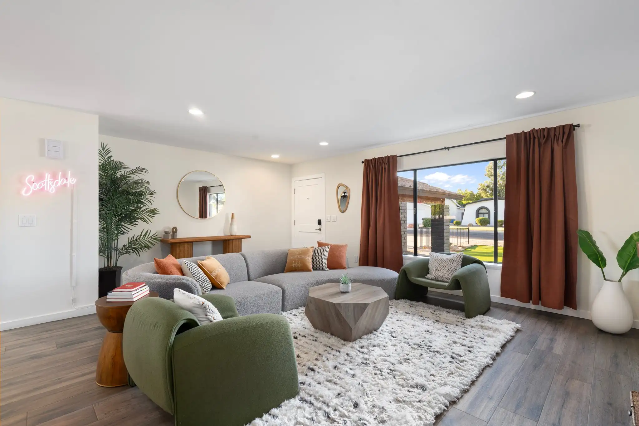 A modern living room with a large gray sectional, two green chairs, and a geometric wood table over a white rug. A neon sign spells "Scottsdale".
