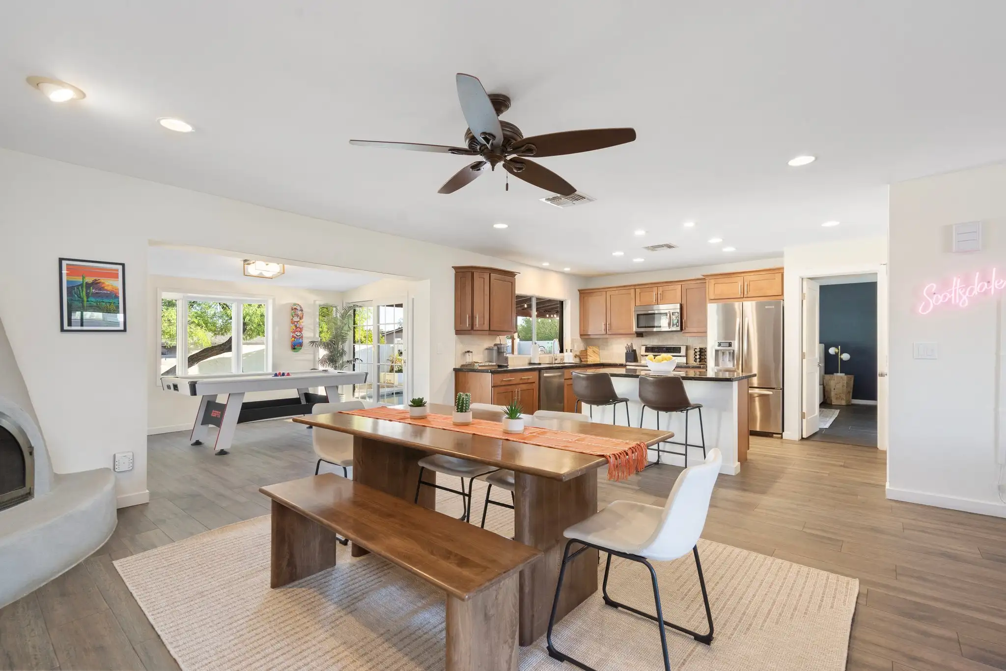 A modern open-concept room features a dining area with a wooden table and bench, leading to a kitchen with wood cabinets and a bar island. An air hockey table is in the back.
