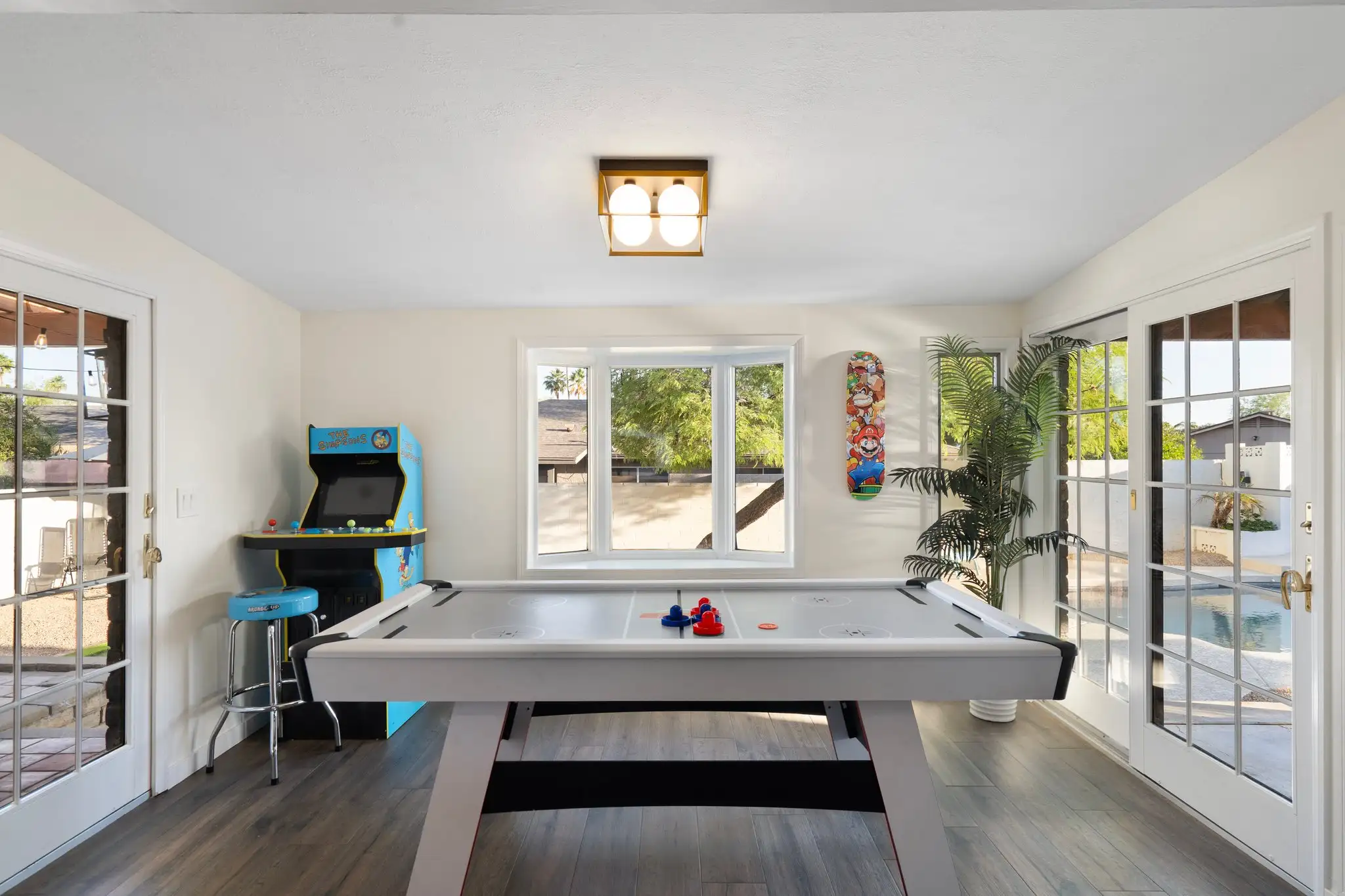 A well-lit game room with an air hockey table, a blue Simpsons arcade machine, and a decorative Mario skateboard. French doors lead out to a backyard pool.
