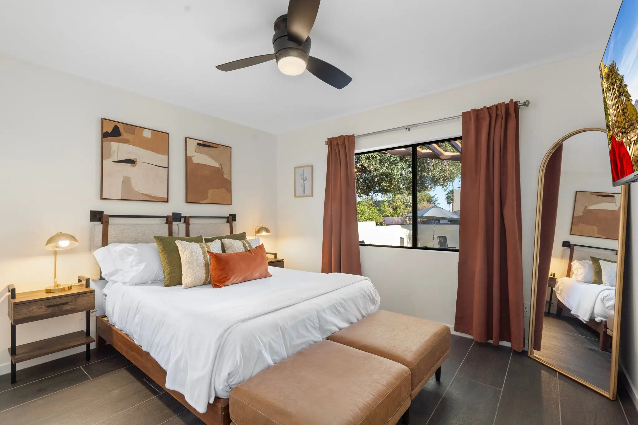 A stylish bedroom with a king bed, warm wood accents, and white walls. Olive green and rust pillows accent the bed, which has two leather ottomans at the foot.