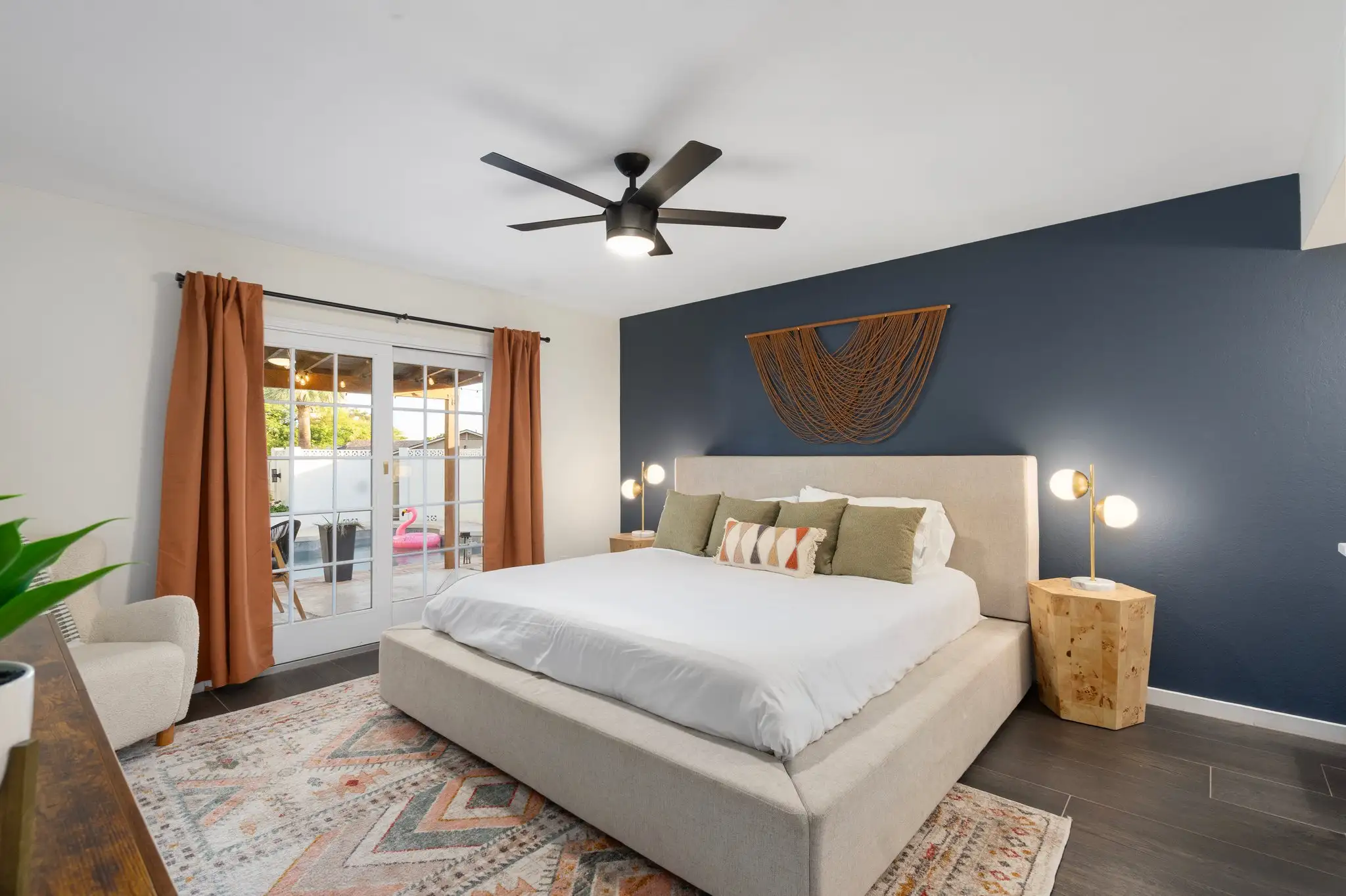 A modern primary bedroom with a large bed centered against a navy wall. Bohemian style decor includes a patterned rug, wood nightstands, and orange curtains framing patio doors.