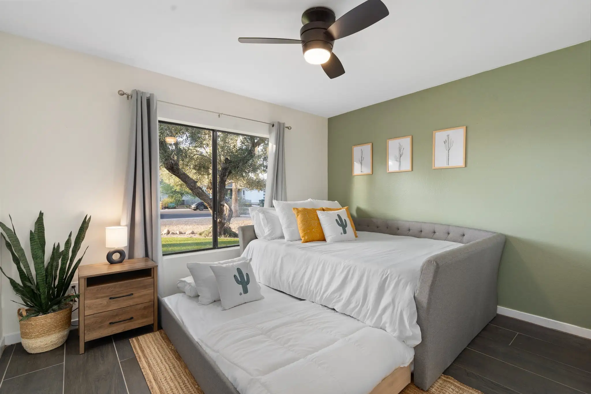 A bright modern guest room featuring a gray daybed with a trundle pulled out and white bedding decorated with cactus-themed pillows. A sage green accent wall and desert plants complete the look.