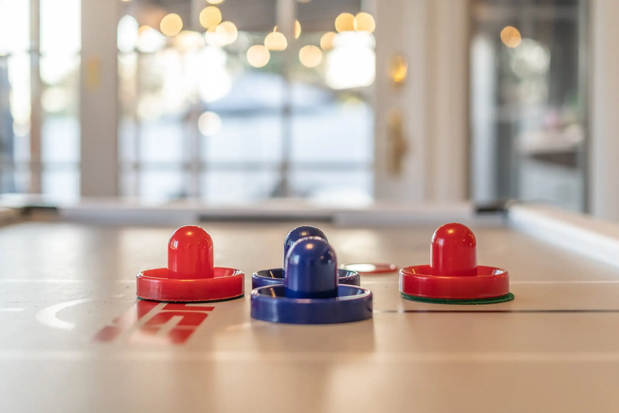 The image shows two red and two blue air hockey mallets resting on a clean white table surface. The background is a bright, blurred interior.