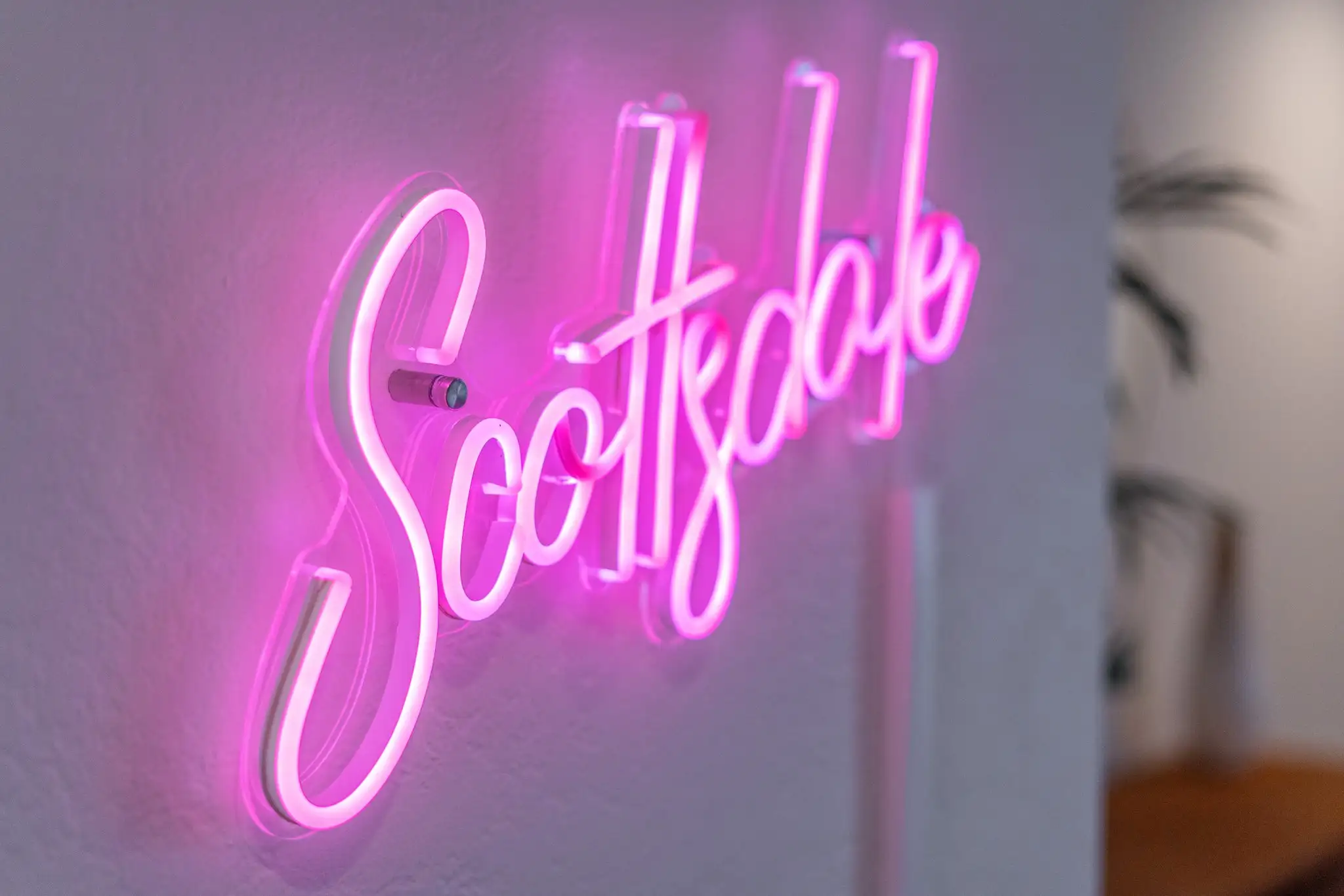 A close-up of a pink neon sign spelling "Scottsdale" in cursive, mounted with standoffs against a white wall.