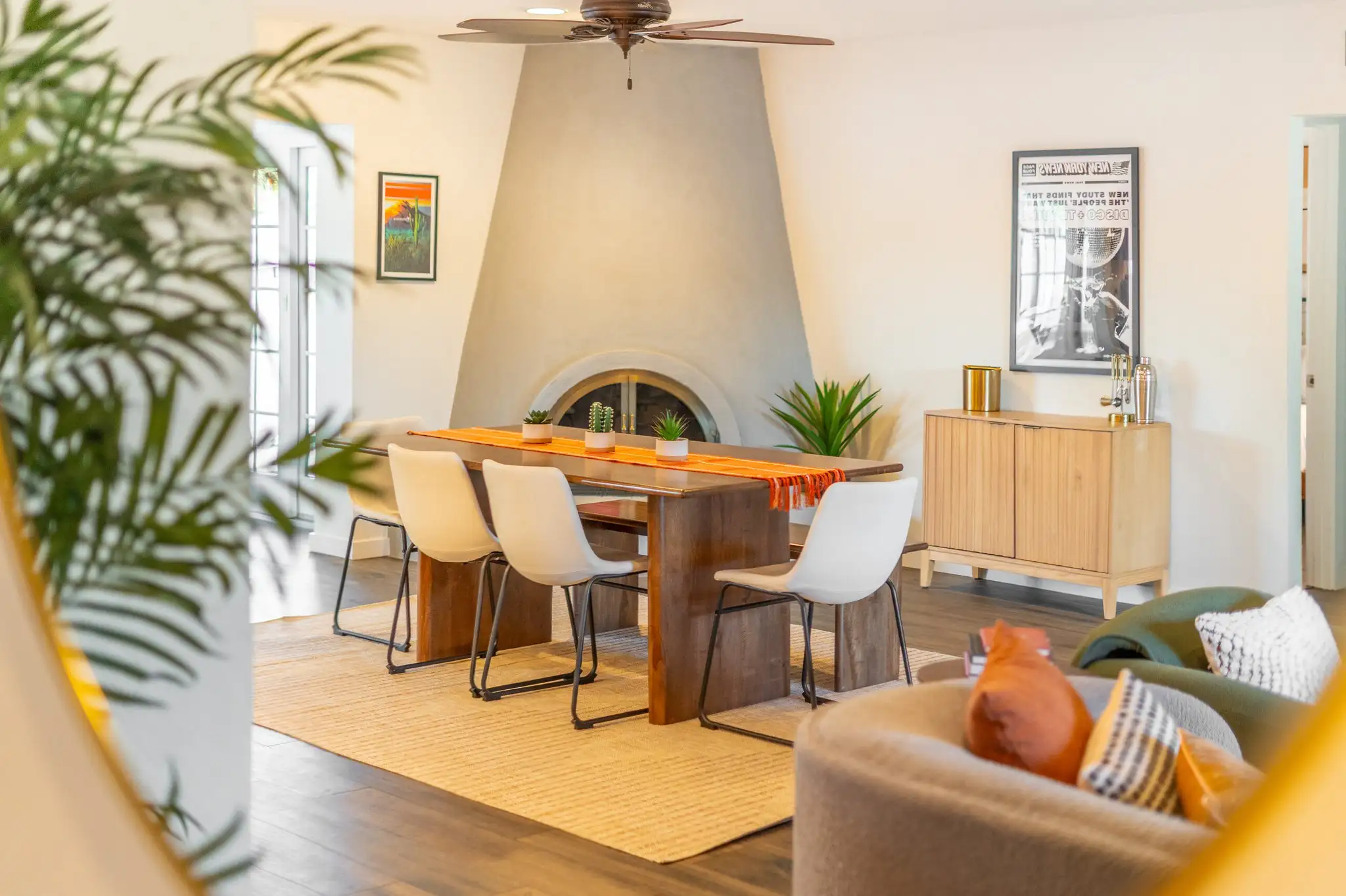 A modern dining area features a wooden table and white chairs, centered beneath a ceiling fan and in front of a large, stucco-style fireplace.