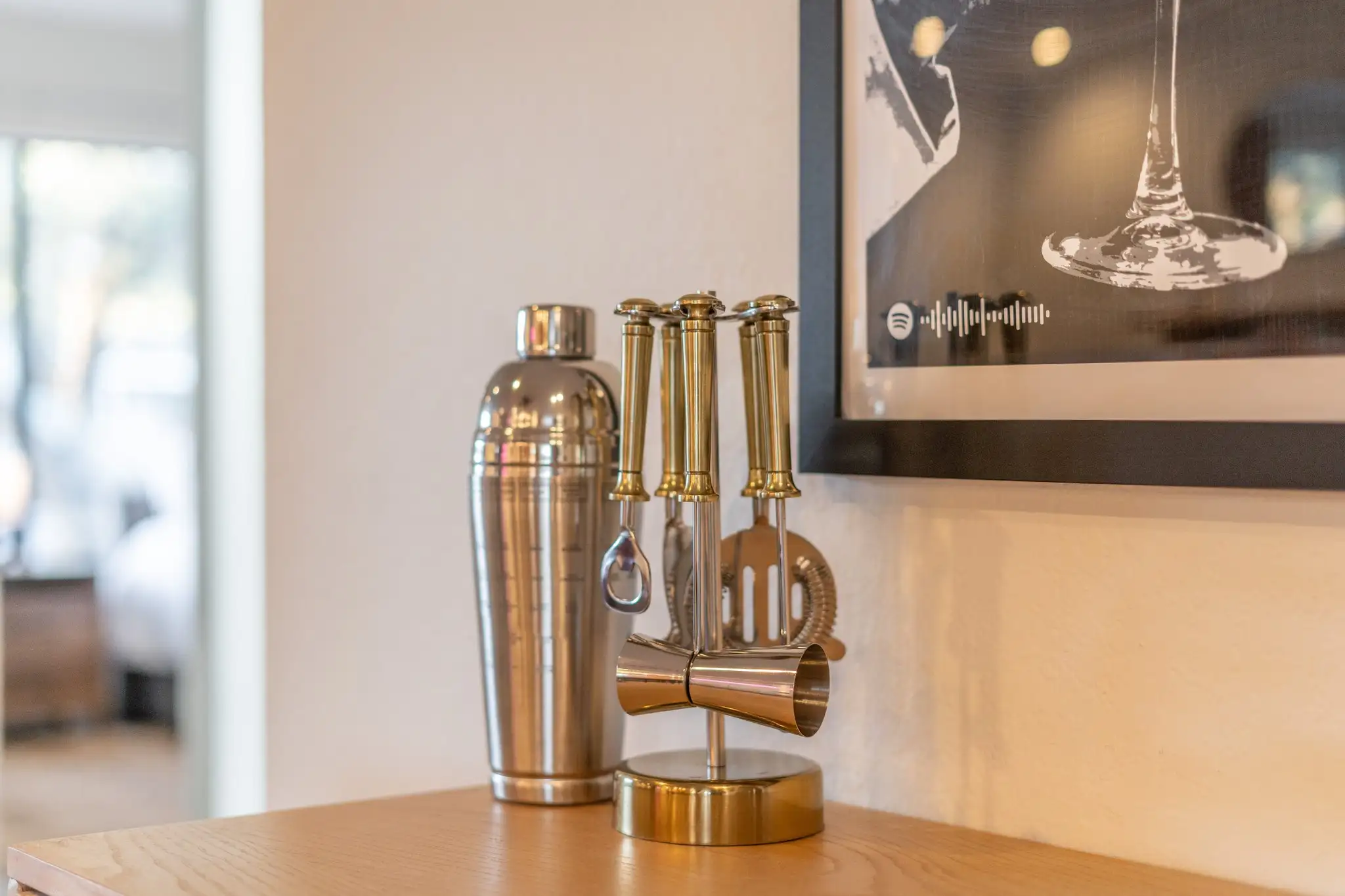 A stainless steel cocktail shaker stands next to a gold bar tool set on a light wood counter, with framed artwork nearby.