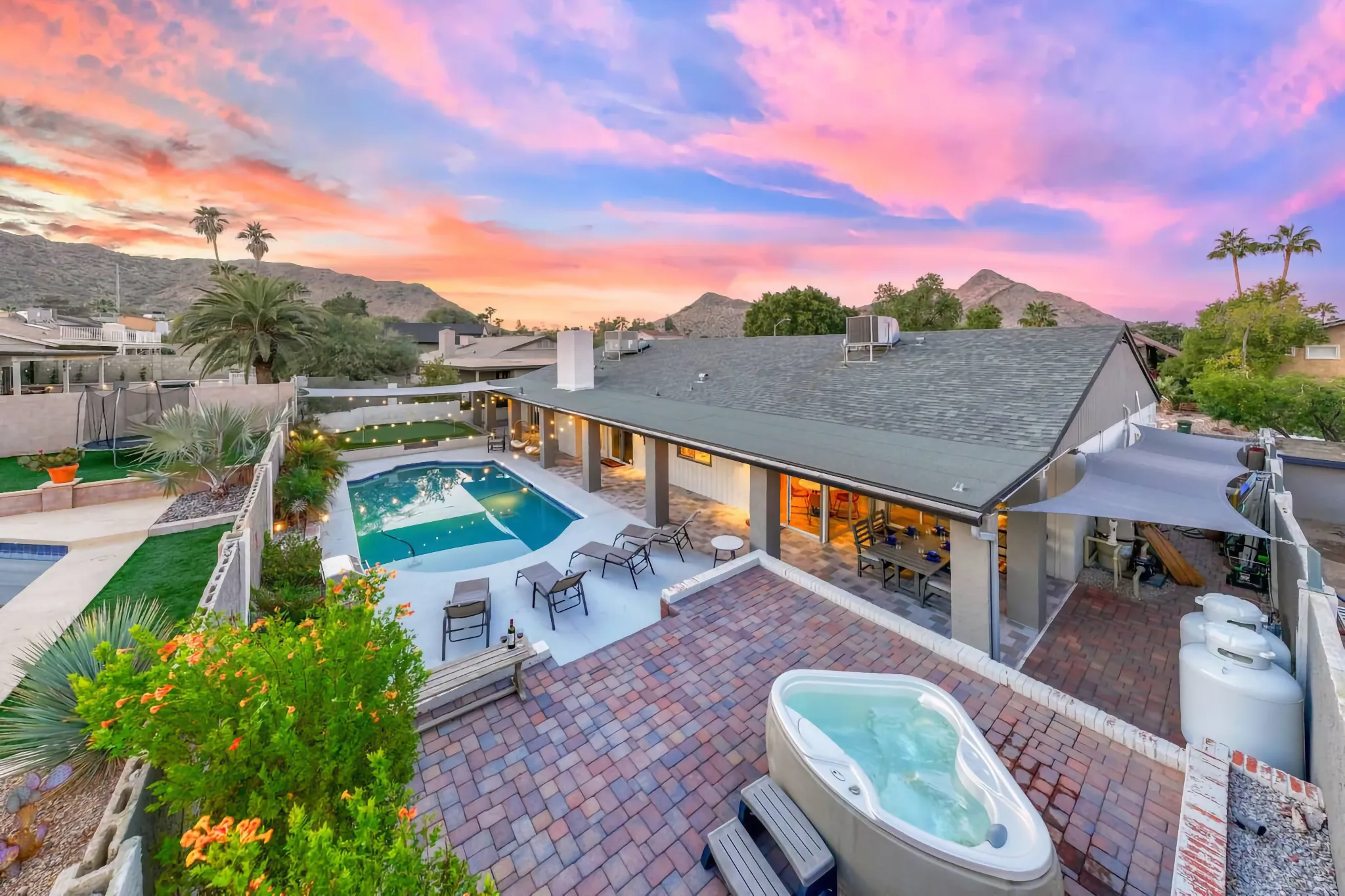 An elevated view of a vibrant residential backyard featuring a pool, hot tub, and patio under a dramatic pink and purple sunset sky over rolling desert hills.