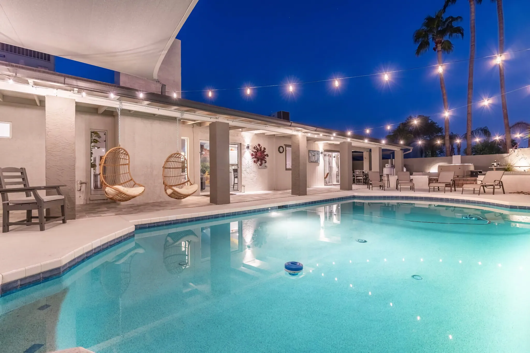 A modern pool area illuminated at night under a deep blue sky. The patio features two hanging wicker chairs and decorative string lights.